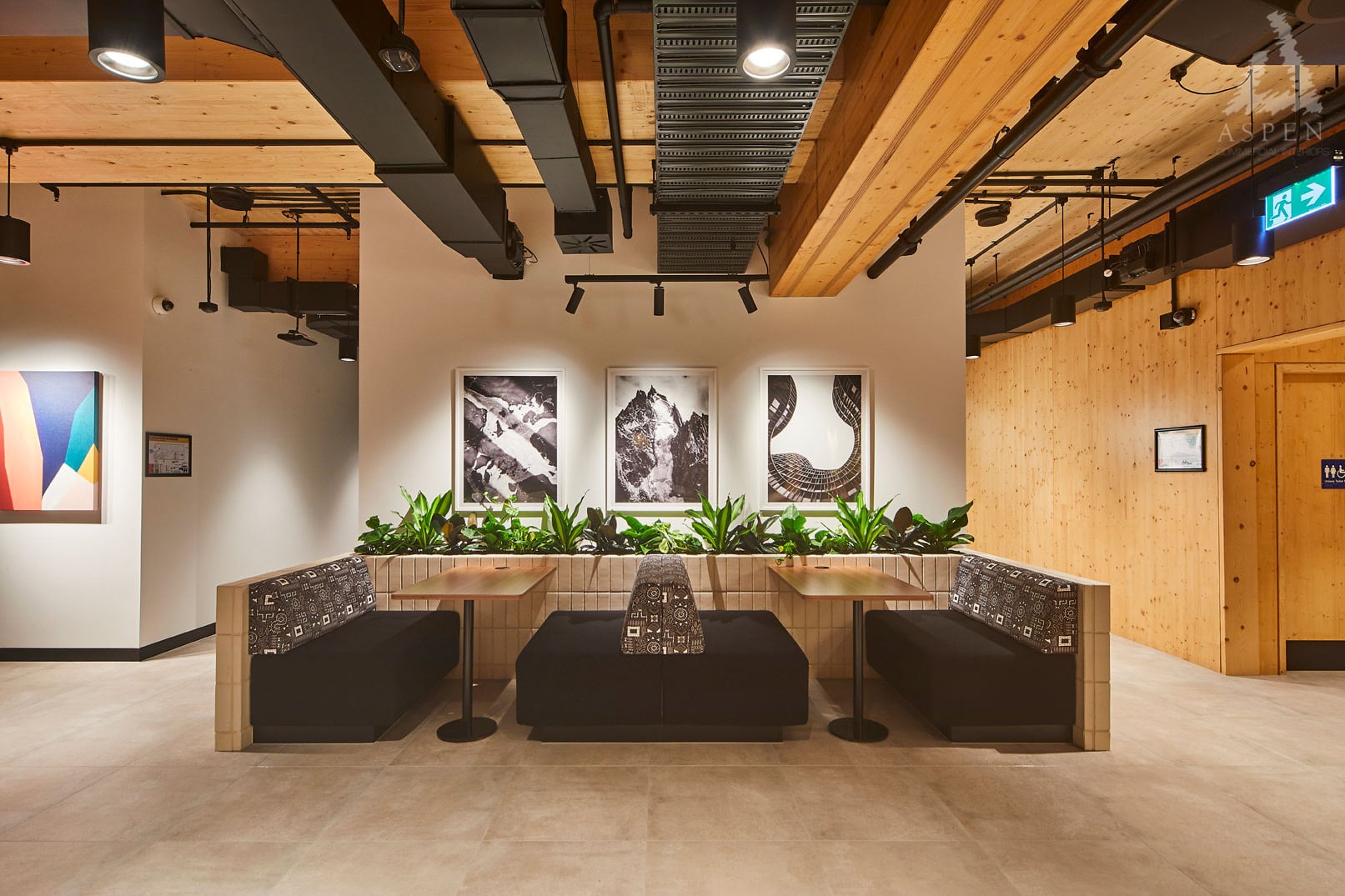 Modern office lounge area featuring black booths, wooden walls, and bright indoor plants under warm lighting.