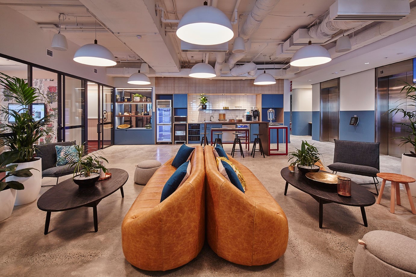 Modern office lounge featuring tan leather sofas, dark coffee tables, indoor plants, and bright pendant lighting.