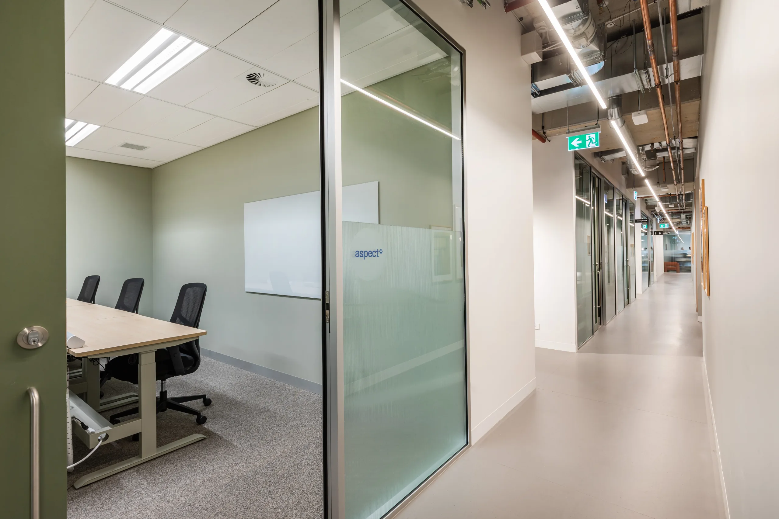 A bright glass-walled meeting room with a large table and chairs sits adjacent to a long office corridor.