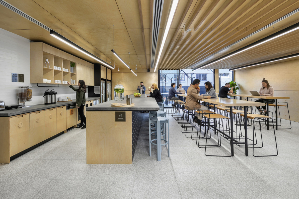 Modern office kitchen and communal dining area with light wood finishes and large windows overlooking the street.