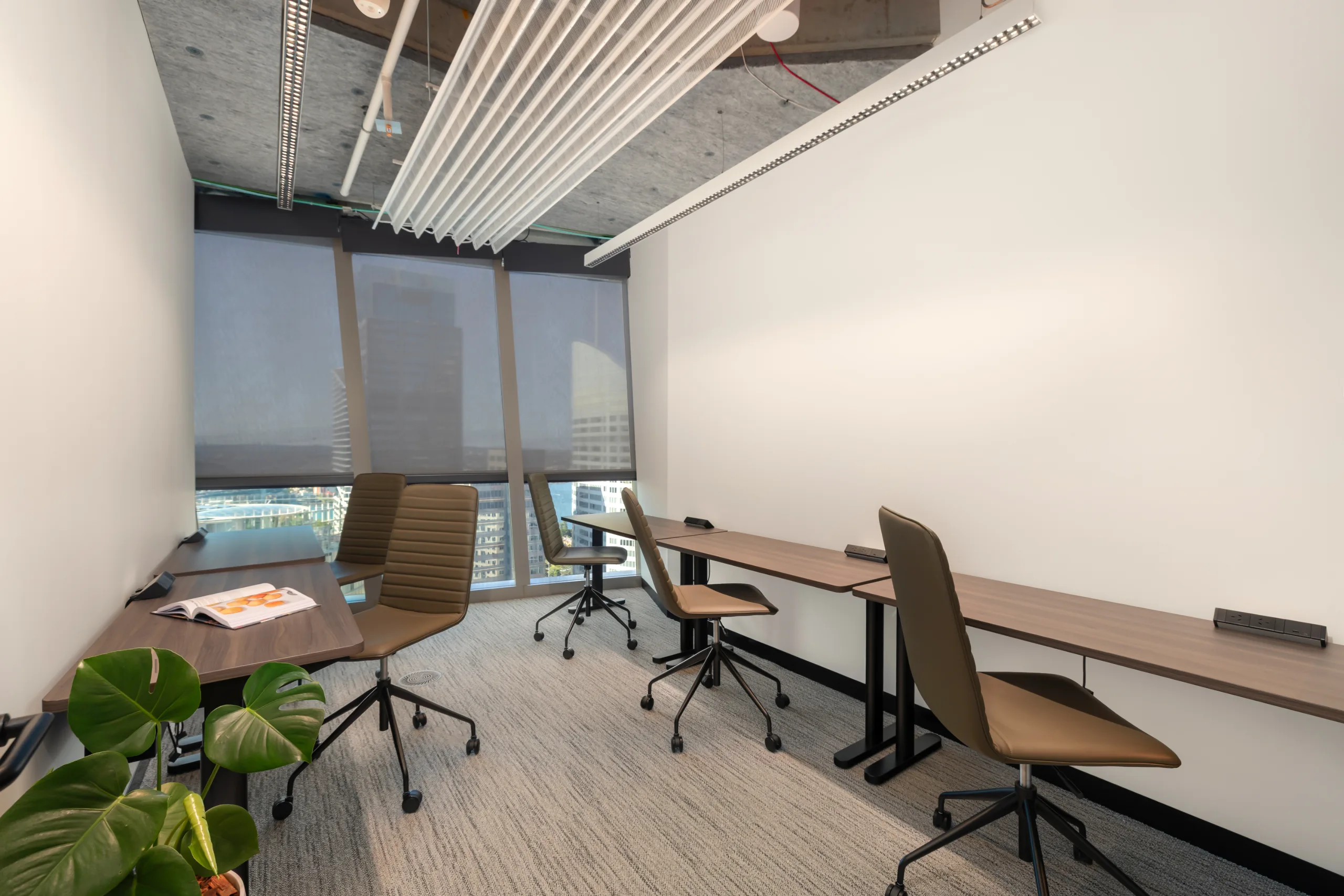 Modern office space with long wooden desks, ergonomic chairs, and large windows overlooking a city skyline.
