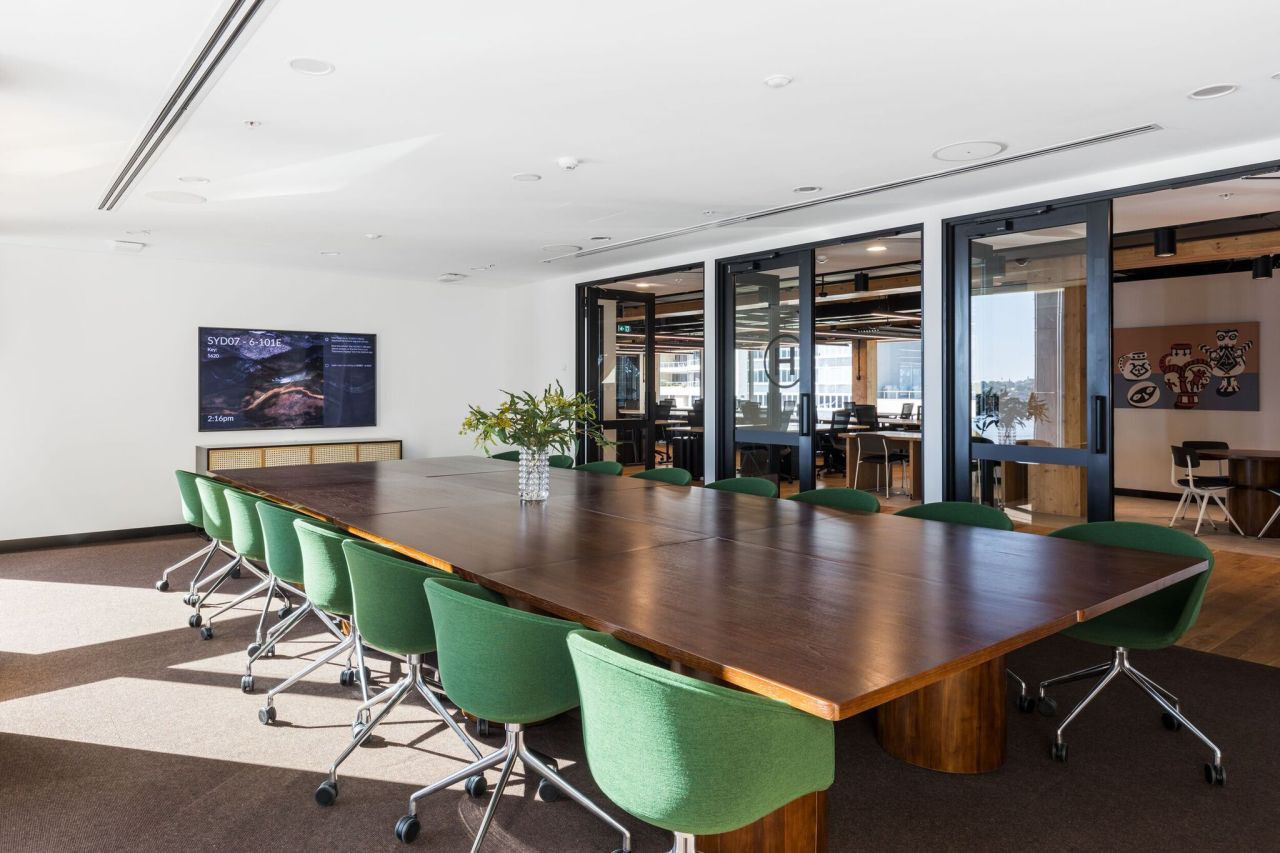 Bright meeting room featuring a long wooden table, green chairs, and large glass doors with natural light.