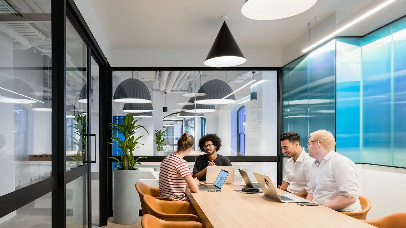Four colleagues sit at a long wooden table in a bright, modern office meeting room.