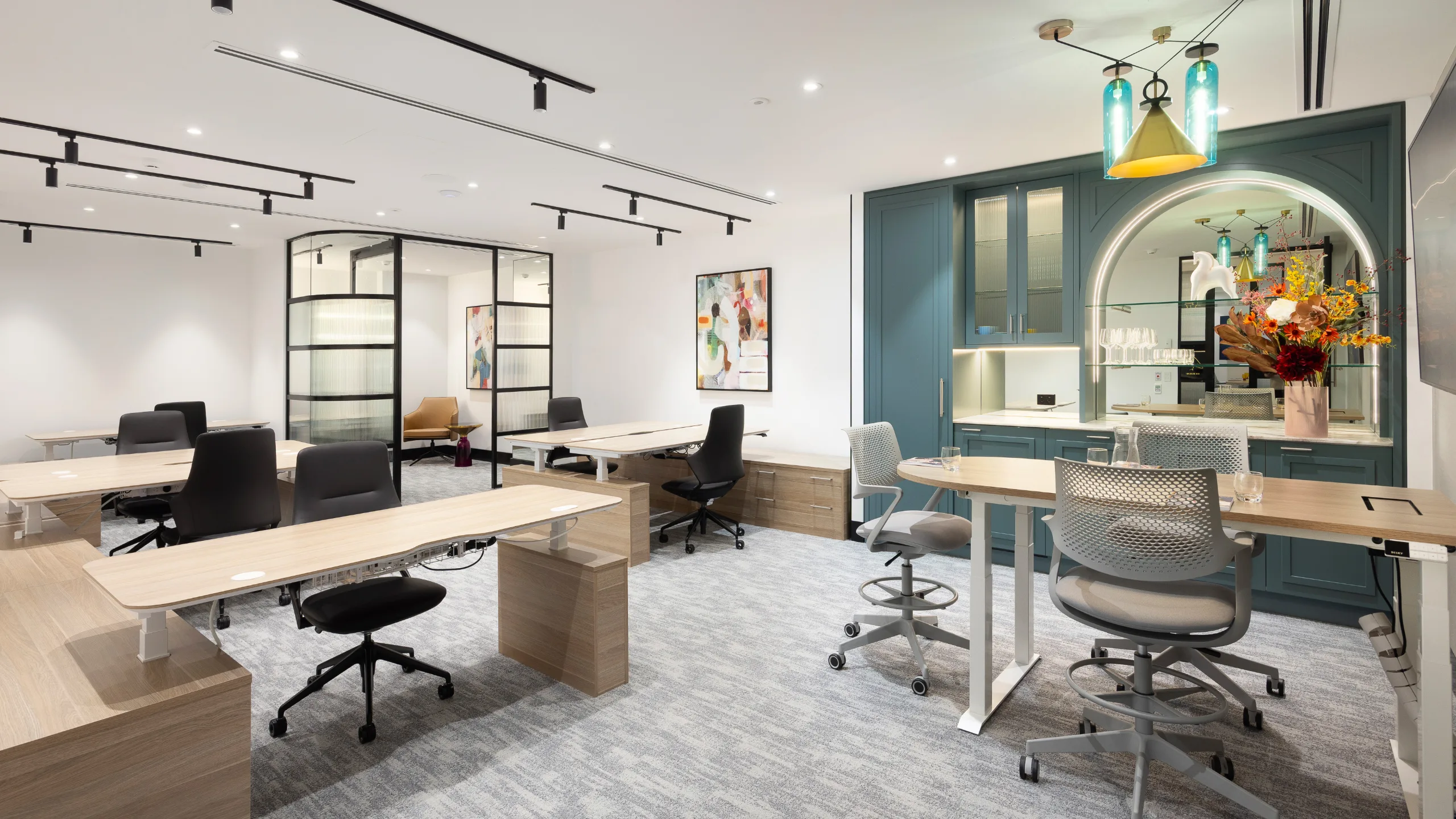 Bright, modern office space featuring wooden desks, ergonomic chairs, and a teal built-in kitchenette area.