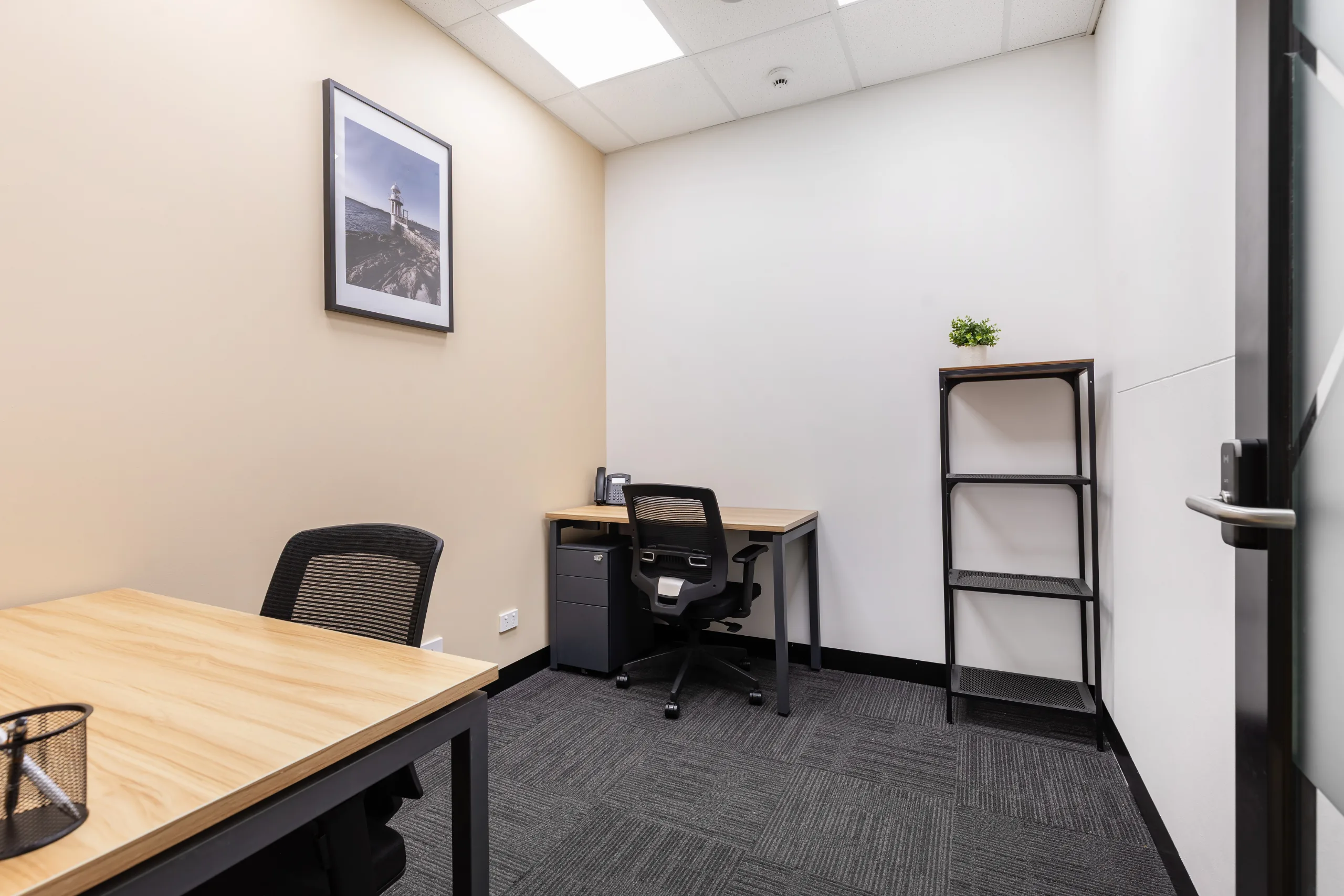 Small private office featuring a wooden desk, black ergonomic chair, and bright artificial lighting.