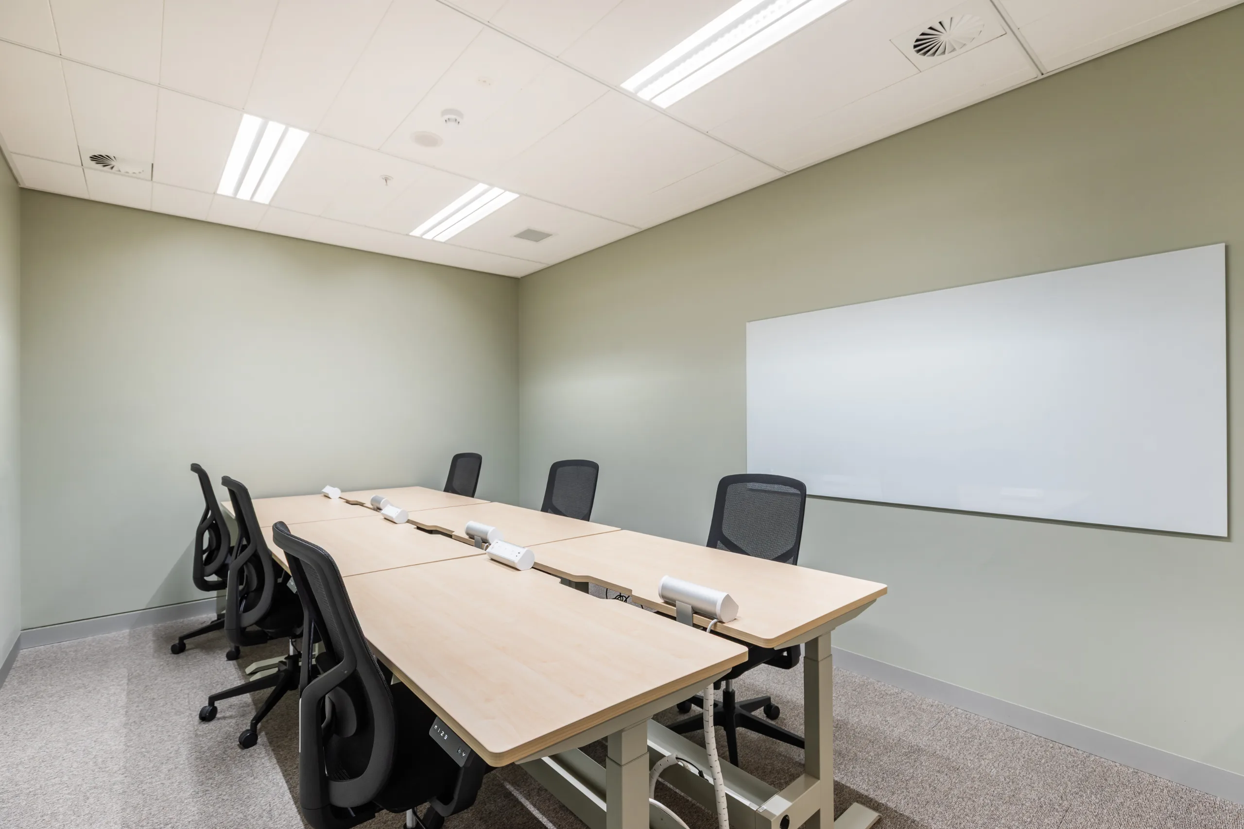 Brightly lit meeting room featuring a long wooden table, black office chairs, and a large whiteboard.