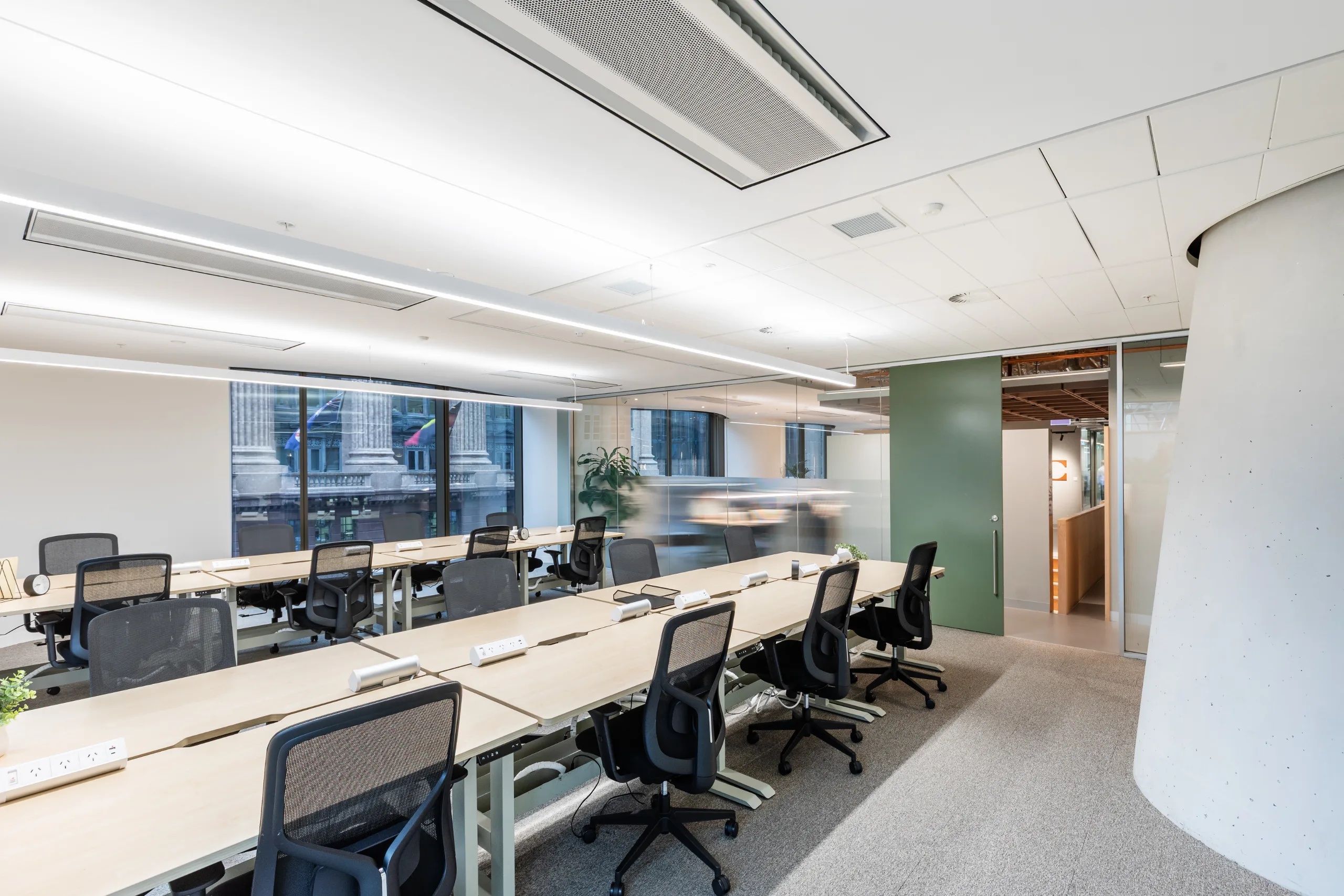 Bright, modern office space with long wooden desks, black ergonomic chairs, and large windows overlooking city buildings.