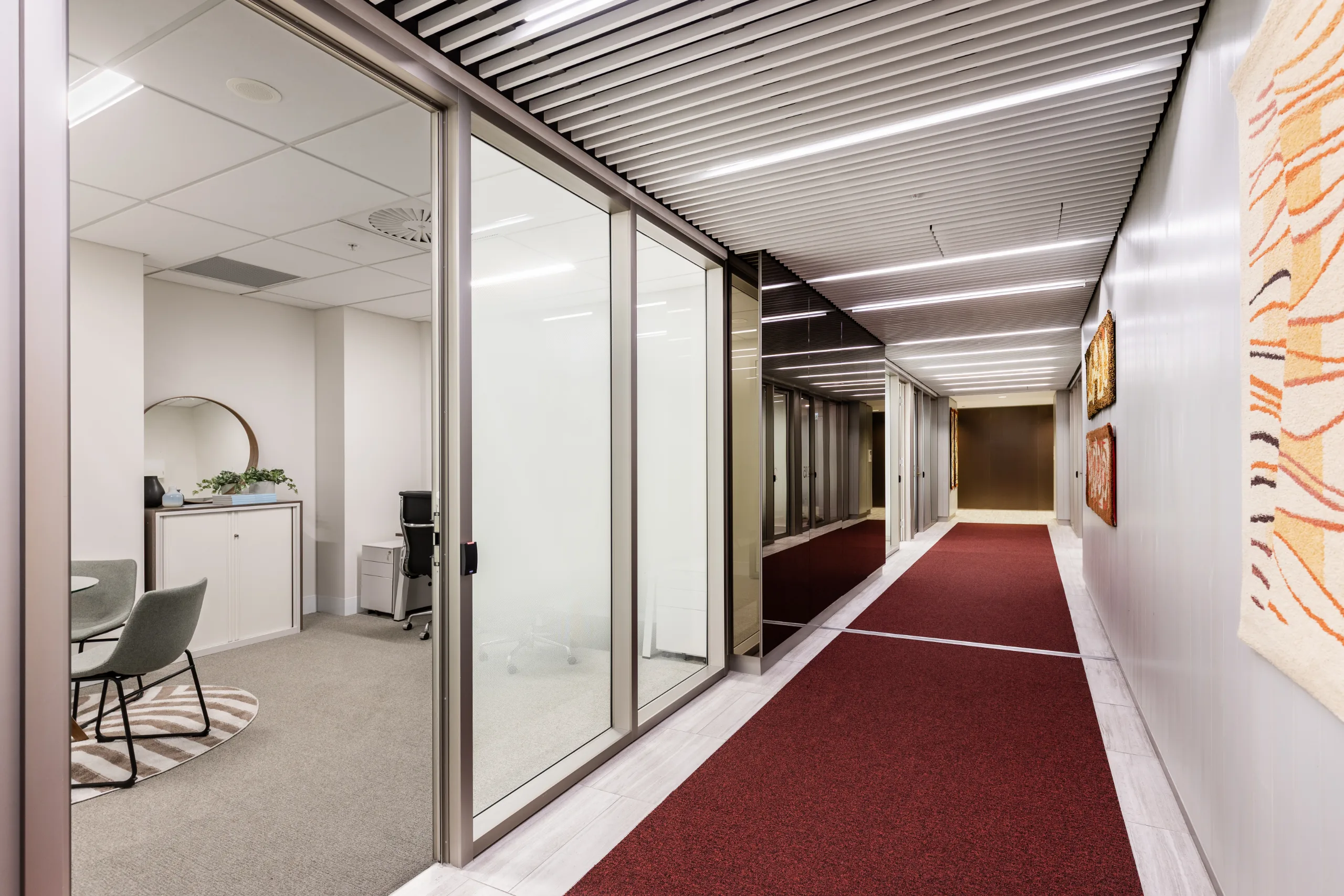 Brightly lit office corridor with frosted glass partitions and a long red carpeted walkway.