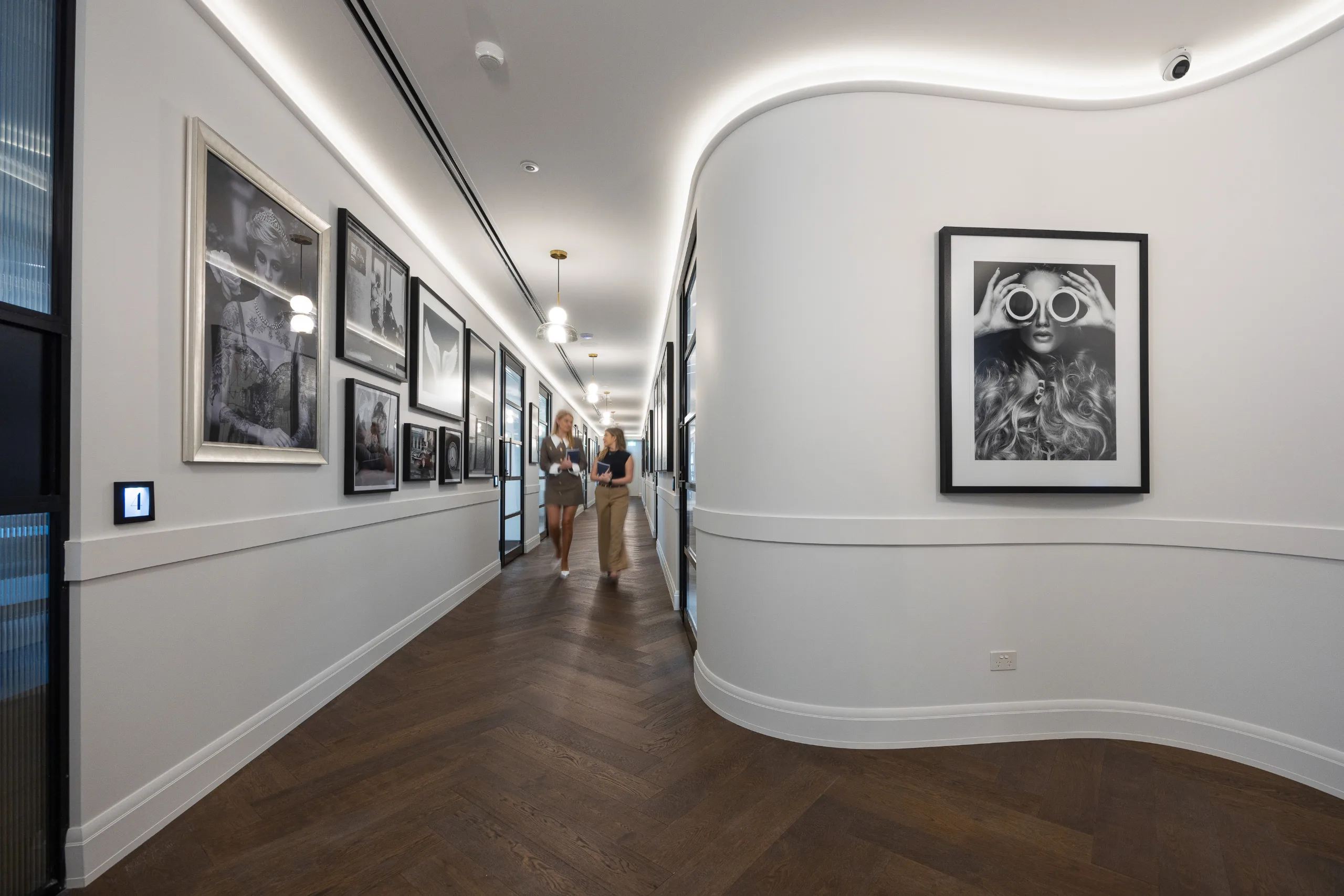 A bright, curved office hallway features dark timber floors, white walls, and framed black and white artwork.