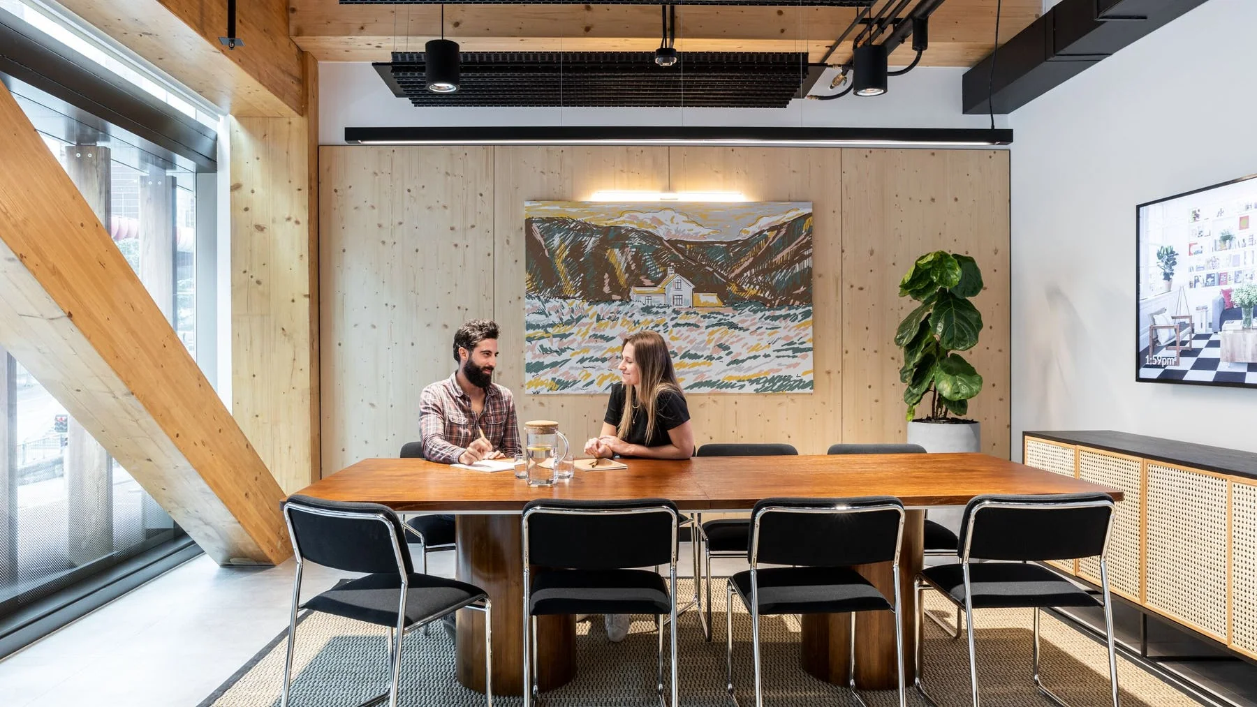 Two people sit at a long wooden meeting table in a bright, modern office with wood-panelled walls.