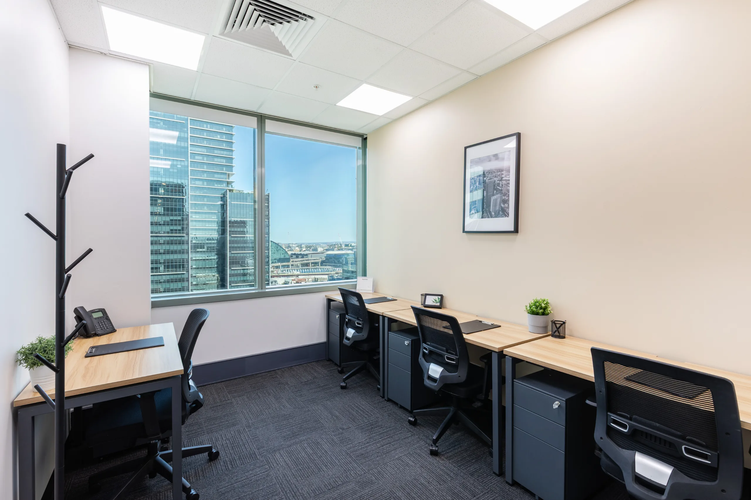Bright office space with wooden desks, black ergonomic chairs, and large windows overlooking city buildings.