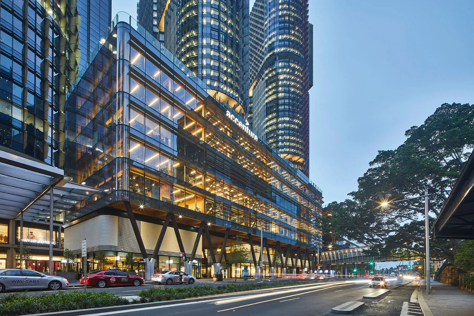 Modern multi-storey glass office building with illuminated interior levels overlooking a city street at dusk.