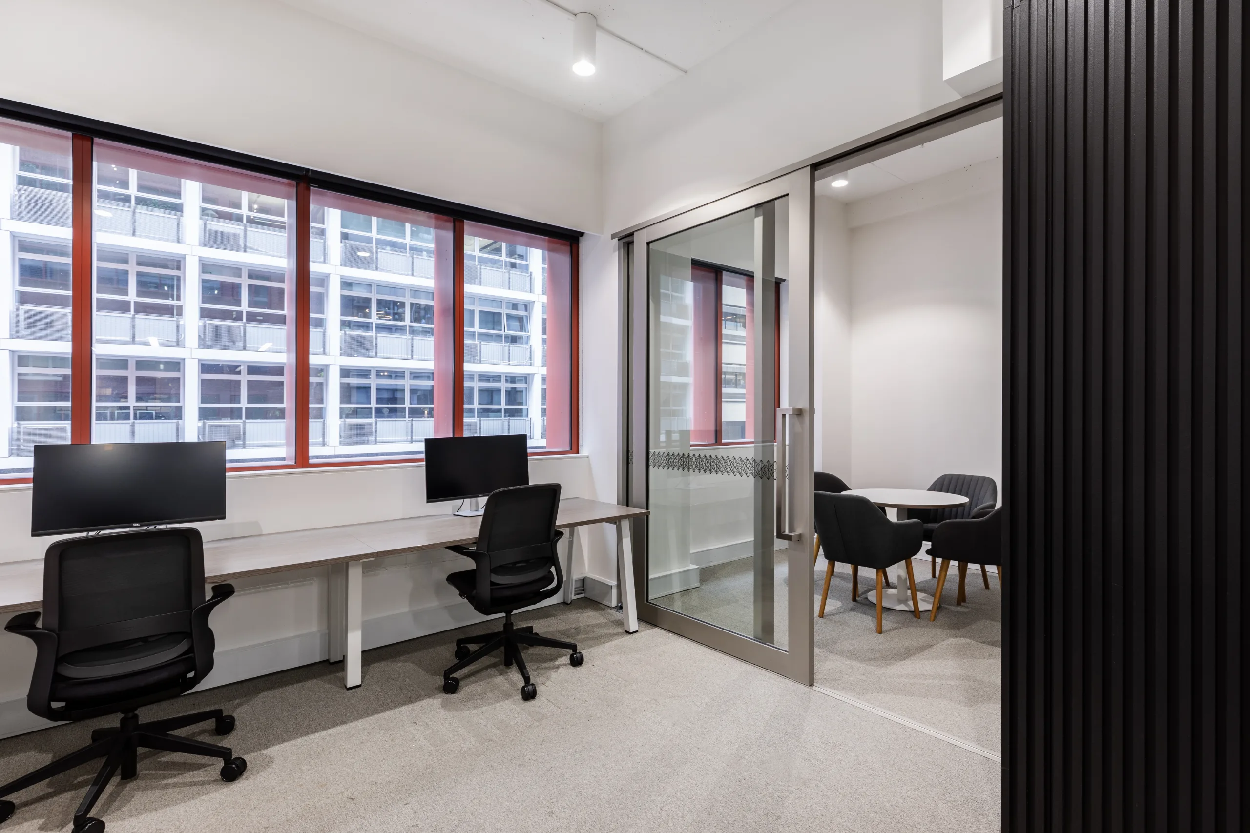Bright office space with white desks, black chairs, and large windows overlooking a nearby building.