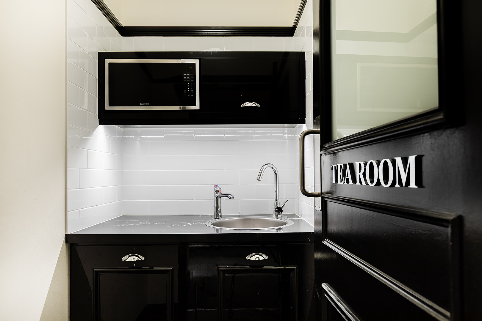 Small tea room featuring a black kitchenette with a microwave, sink, and white tiled walls.