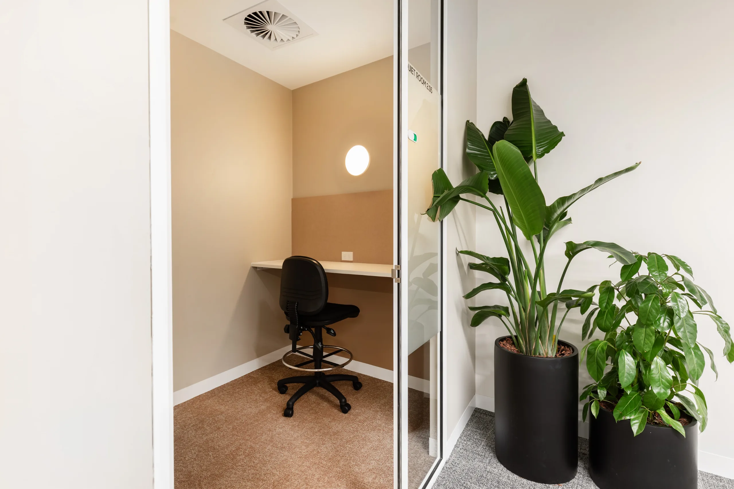 Small private office with a desk, black chair, and warm lighting inside a glass-walled room.