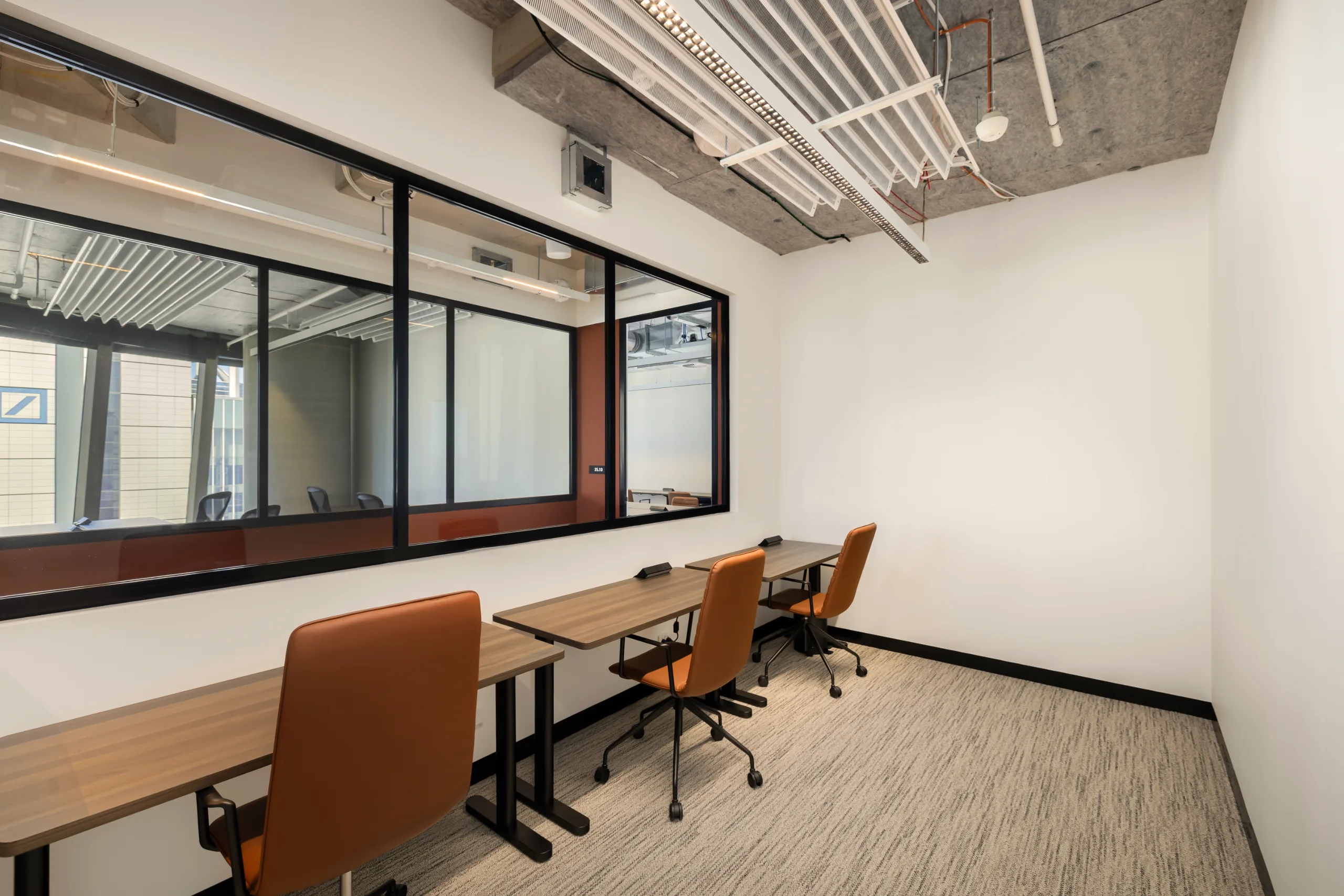 Bright office room with wooden desks, orange chairs, and a large glass partition overlooking another workspace.