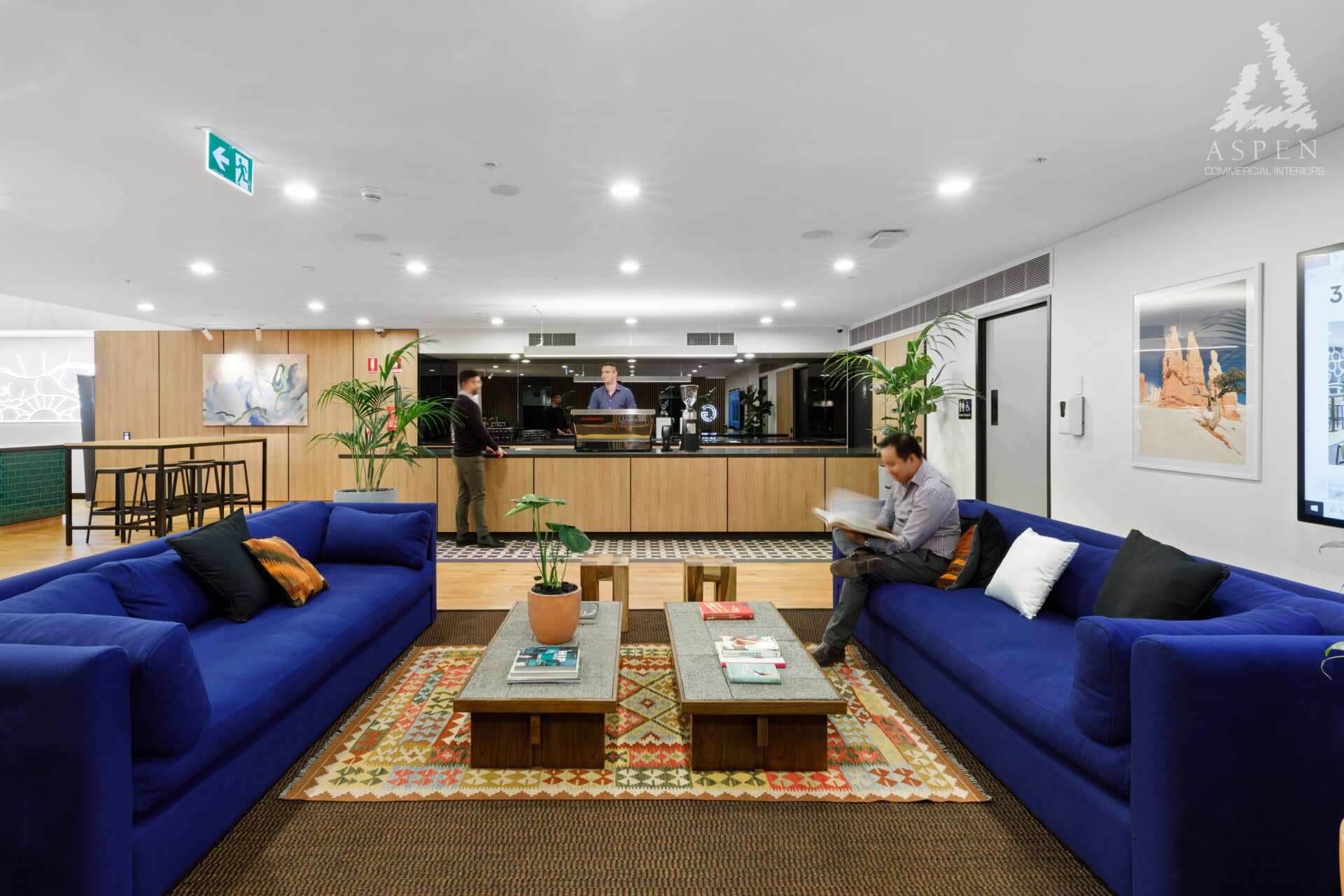 Modern office lounge featuring blue sofas, wooden coffee tables, and a bright kitchenette area in the background.