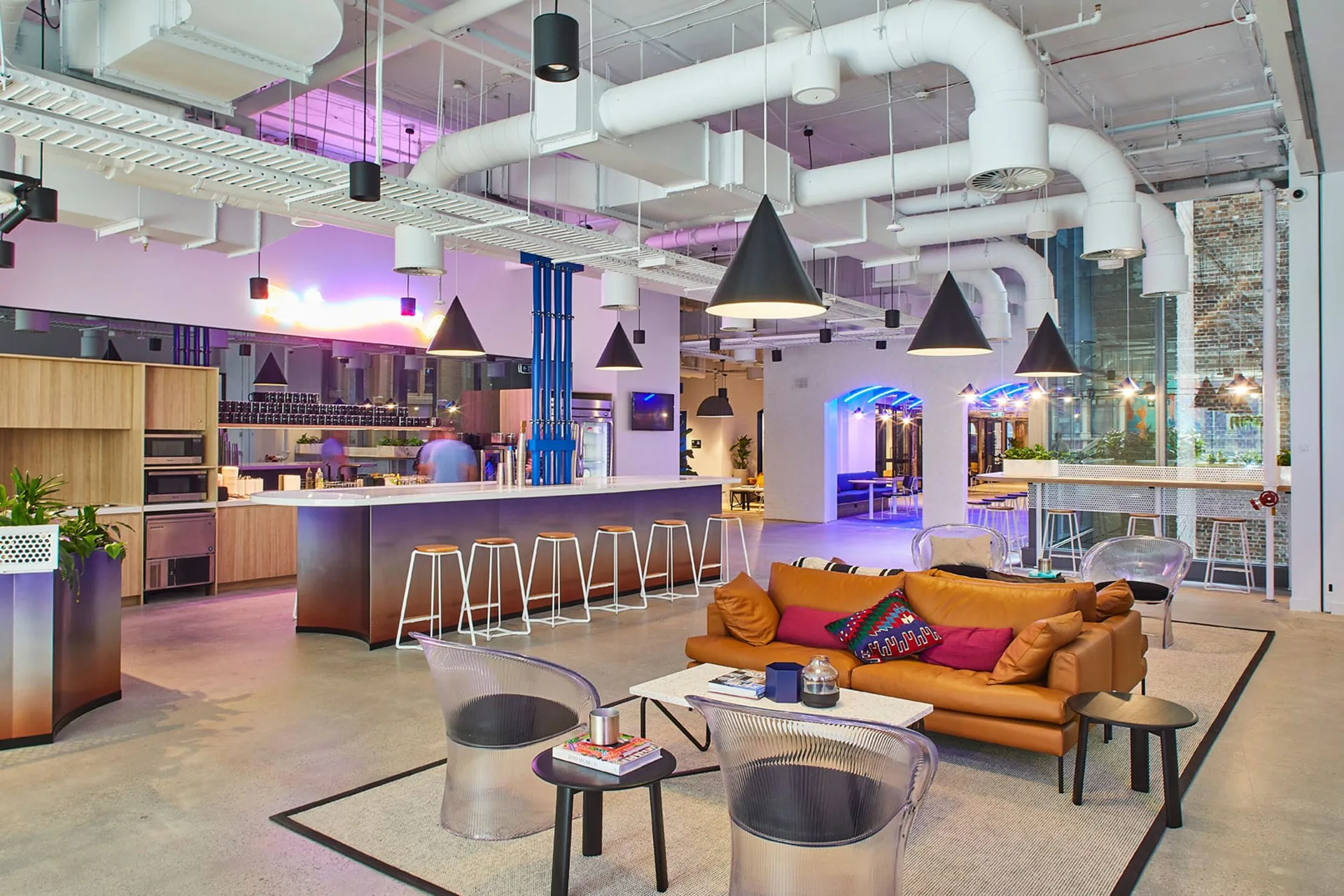 Modern open-plan office lounge featuring a leather sofa, bar seating, and bright industrial lighting.