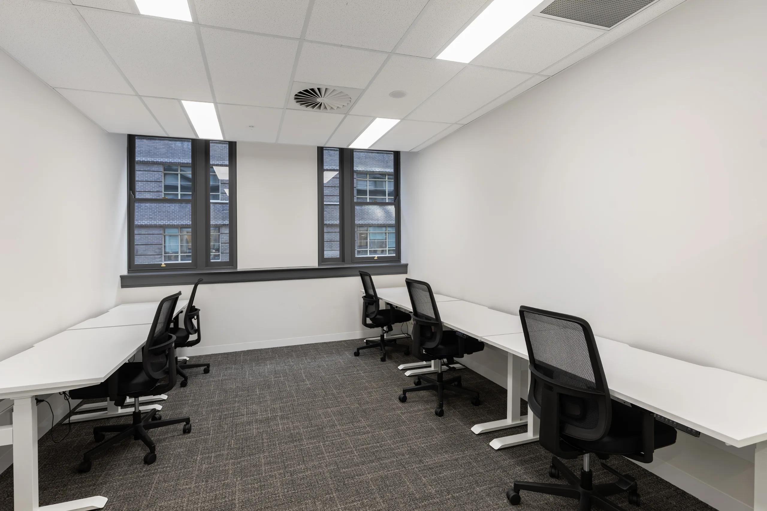Bright office room with white desks, black ergonomic chairs, and large windows overlooking nearby buildings.