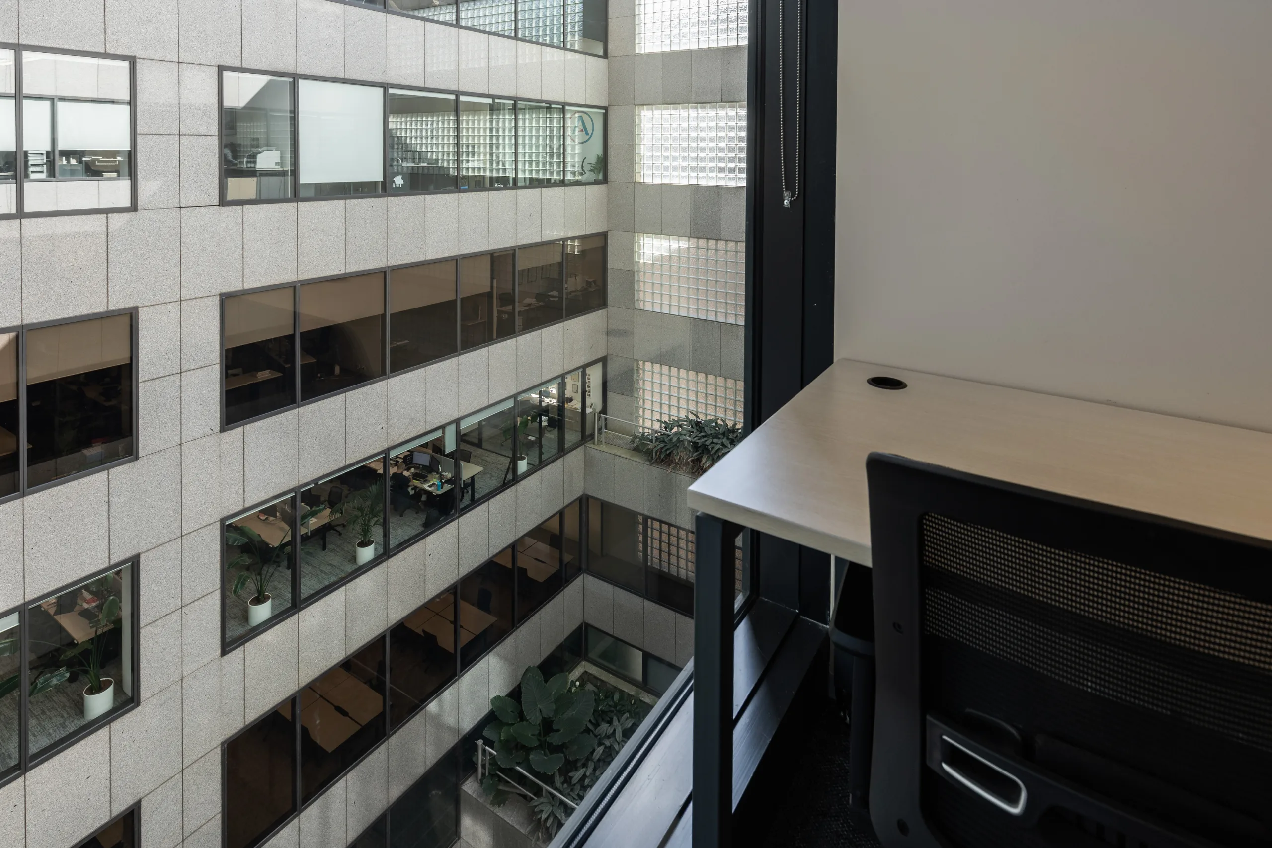 Modern office desk by a large window overlooking a multi-level internal courtyard with glass-walled workspaces.