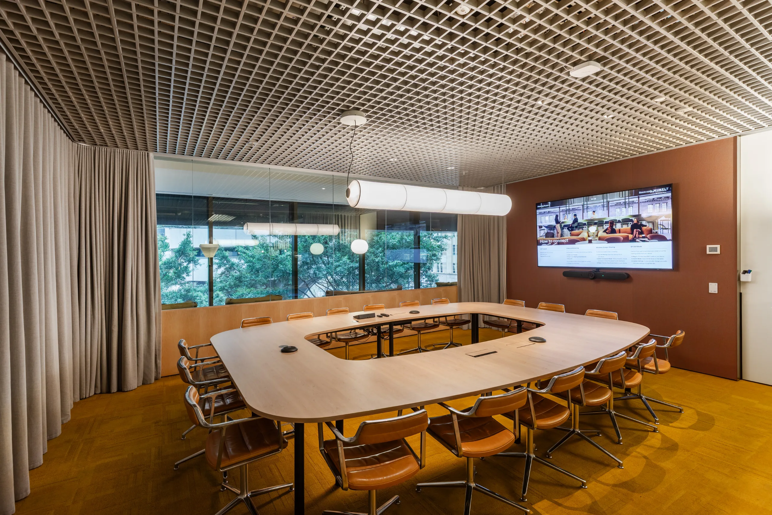 Modern conference room with a large U-shaped wooden table, leather chairs, and large windows overlooking green trees.
