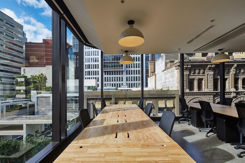 Bright office space featuring a long wooden meeting table and large windows with city views.