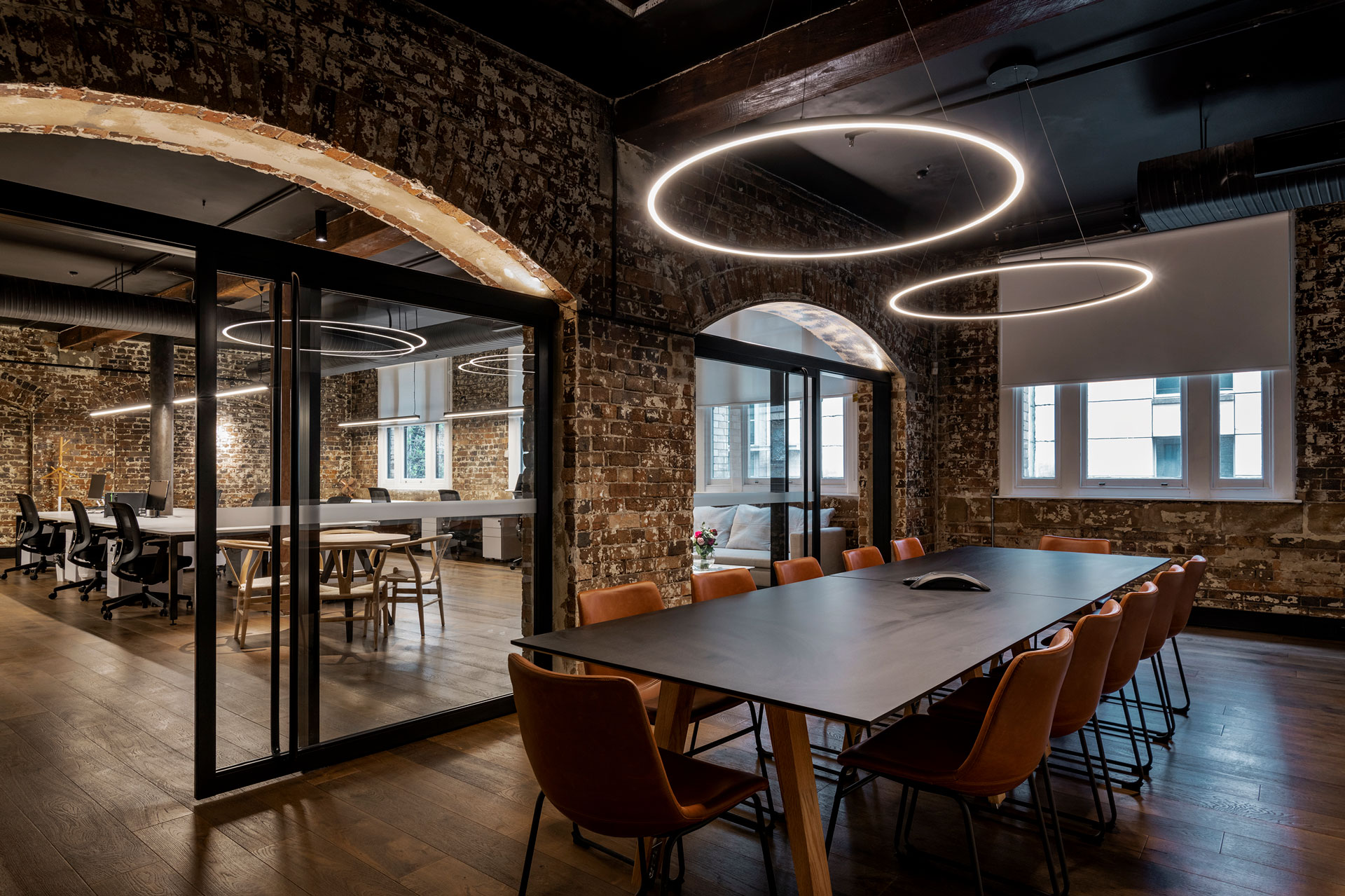 Modern office space featuring a long meeting table, brick walls, and large windows with bright natural light.