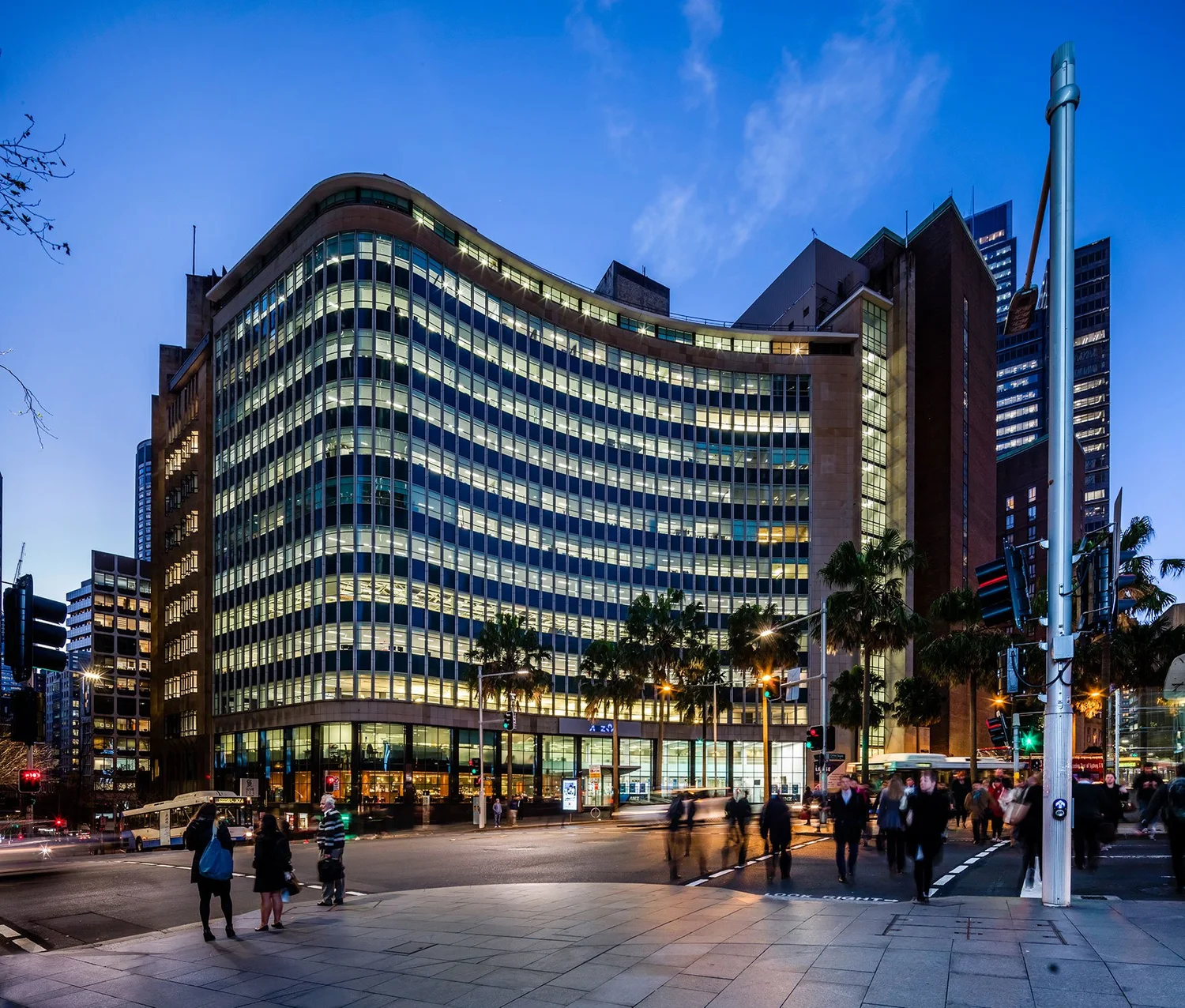 A large, multi-storey curved office building with illuminated windows stands above a busy city street at dusk.