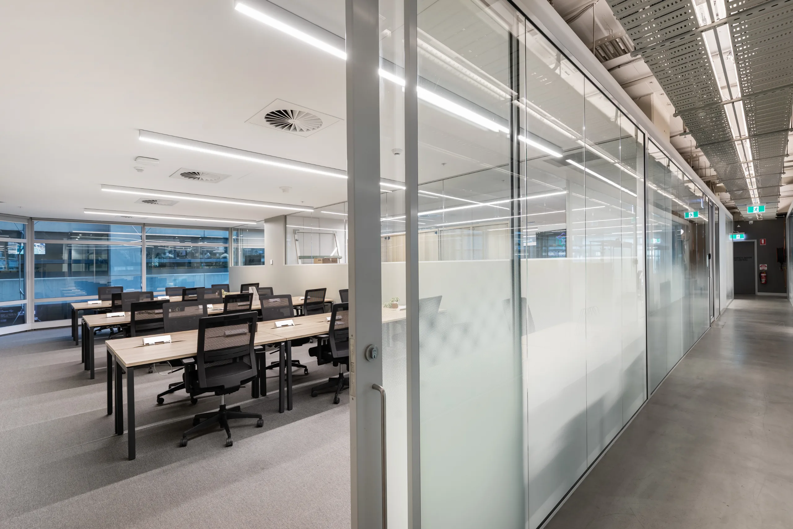 Bright, modern open-plan office featuring rows of desks, black chairs, and large glass partitions.