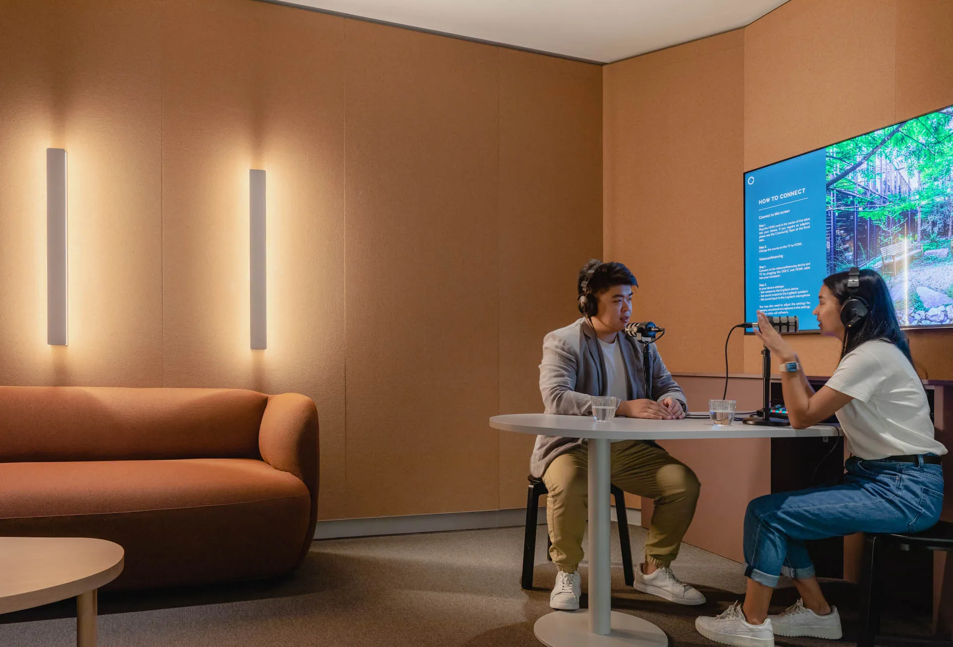 Two people sit at a small table in a modern office lounge with warm lighting and a large screen.