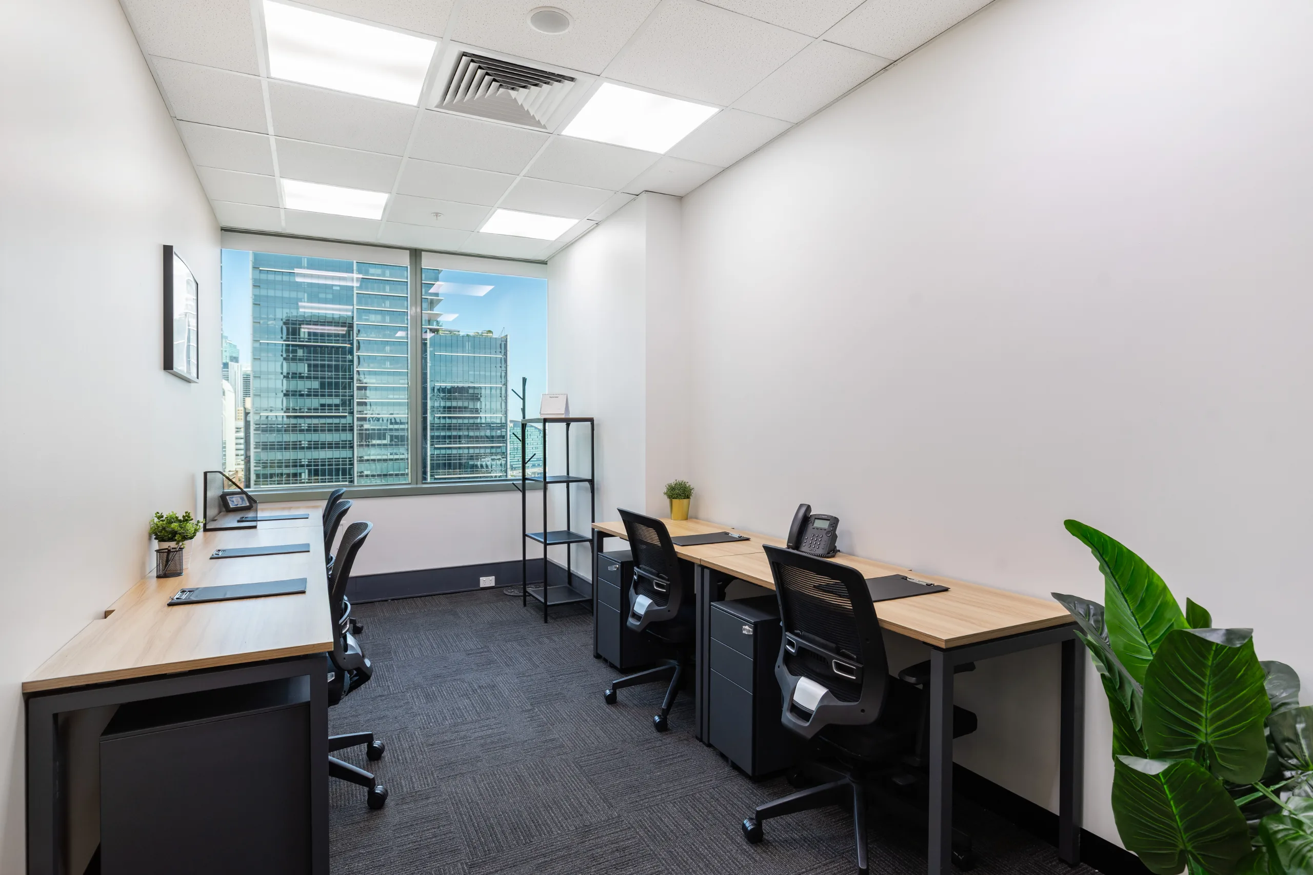 Bright office space featuring two wooden desks, black chairs, and large windows with city views.