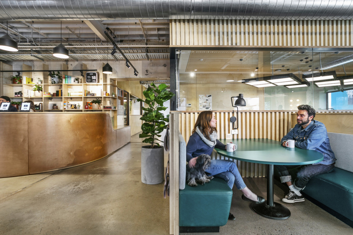 Modern office lounge with a curved wooden reception desk, green plants, and people sitting at a round table.