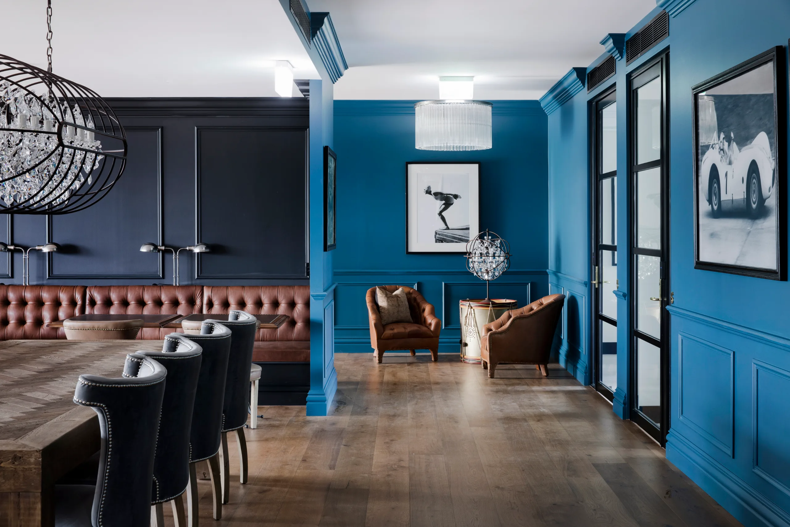 Modern office lounge with blue walls, leather armchairs, wooden floors, and large windows with natural light.