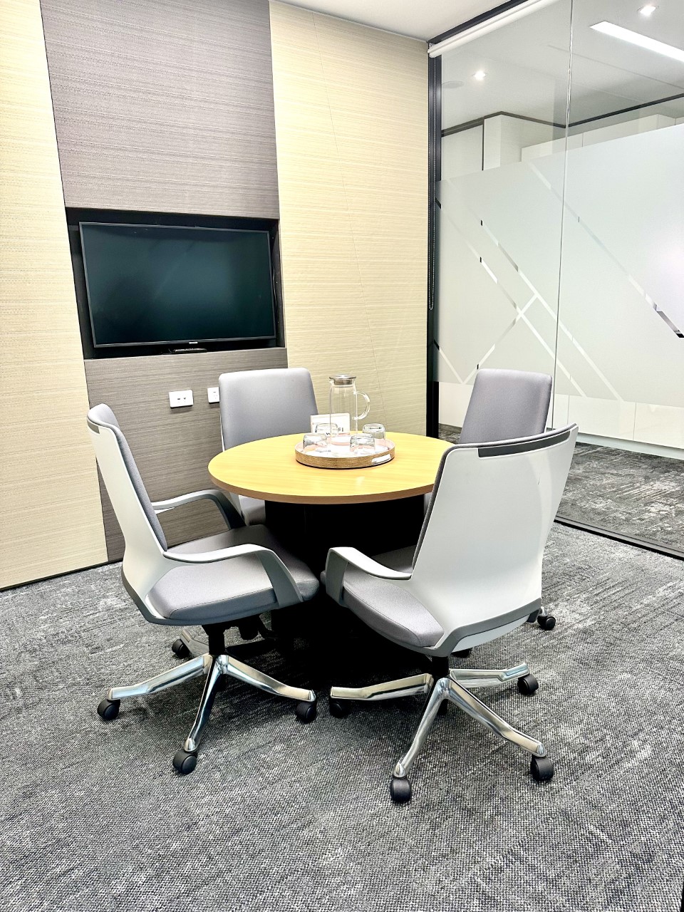 Small meeting room featuring a round wooden table, four grey chairs, and a wall-mounted television screen.