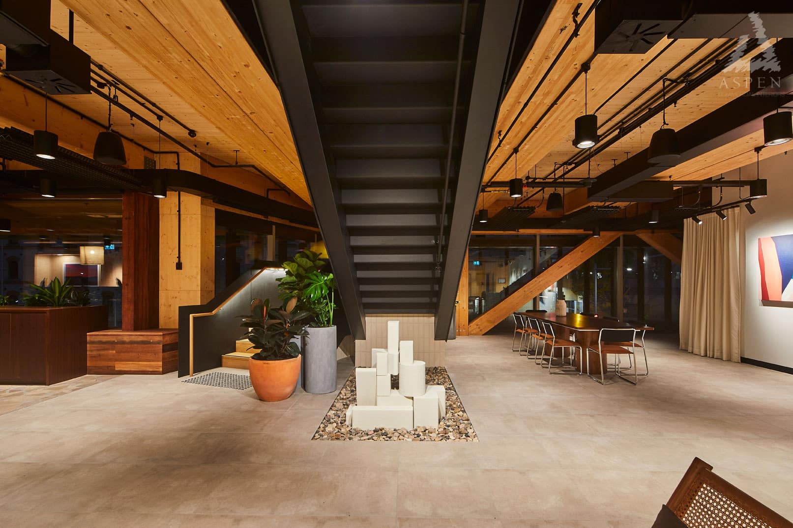 Modern open-plan office space featuring a central black staircase, wooden ceilings, and large windows with natural light.