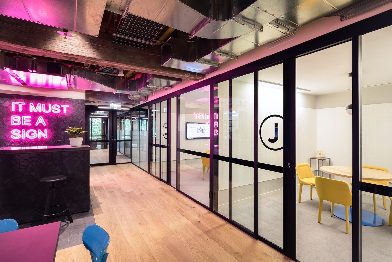 Modern office corridor with glass-walled meeting rooms, wooden flooring, and a bright pink neon sign.