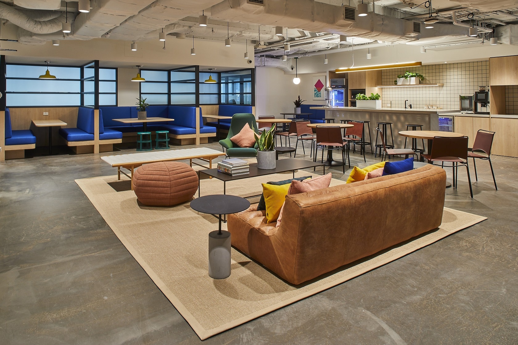 Modern open-plan office lounge featuring comfortable seating, communal tables, and a bright kitchenette area.