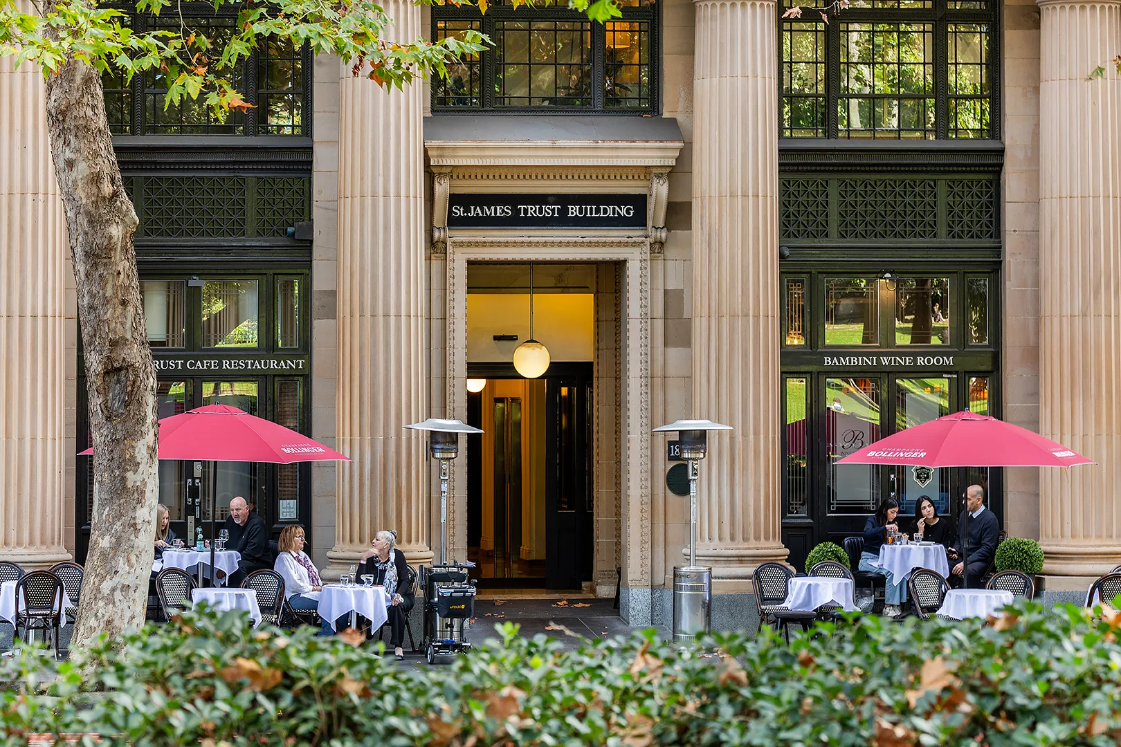 Outdoor cafe seating with red umbrellas sits in front of the St James Trust Building's stone facade.