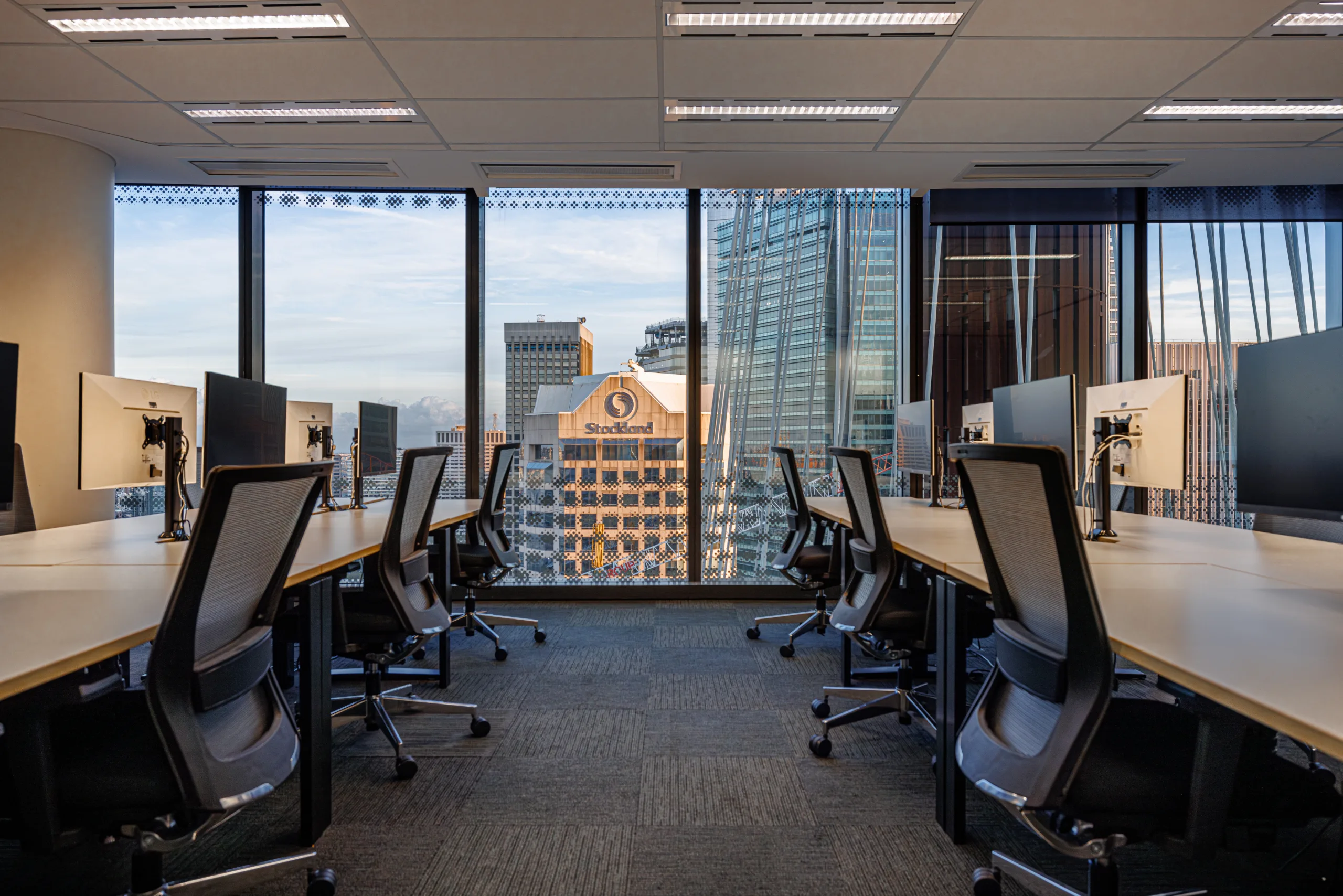 Modern office space with long desks, computer monitors, and large windows overlooking a city skyline.