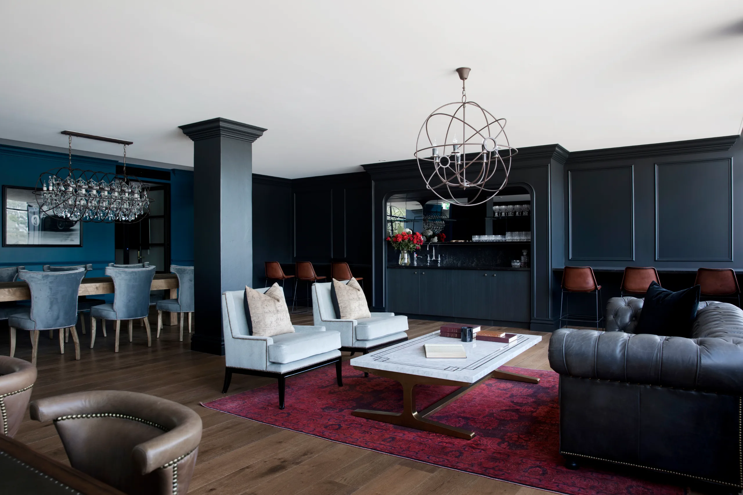 Modern lounge area with dark paneled walls, plush armchairs, a marble coffee table, and a large spherical chandelier.