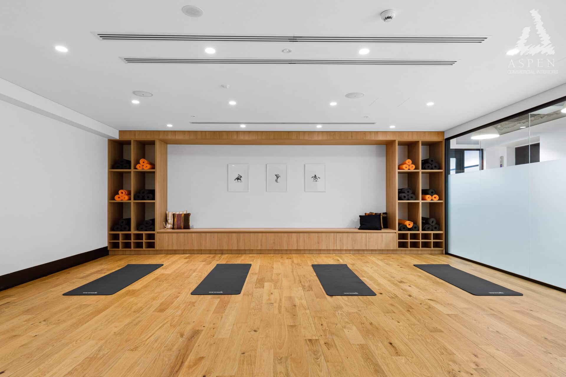 Bright yoga studio with wooden floors, black mats, and built-in timber shelving against a white wall.