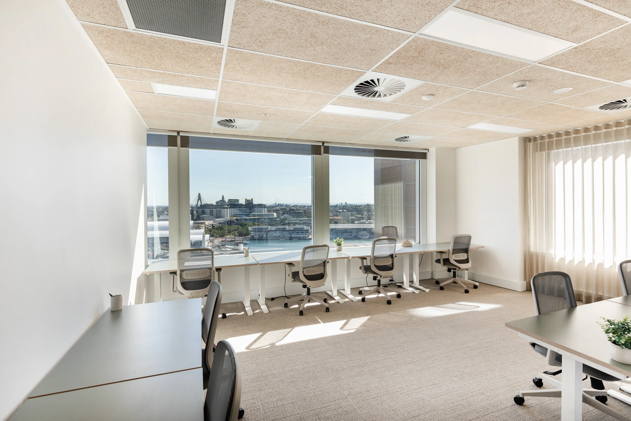 Bright, modern office space with long white desks, ergonomic chairs, and large windows overlooking a city harbour.