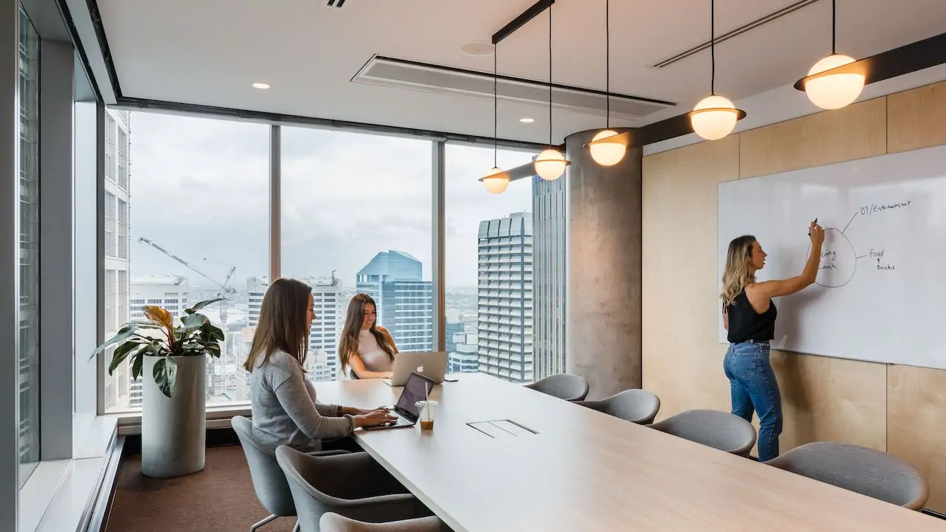 Bright meeting room with a long wooden table, large windows overlooking city skyscrapers, and a whiteboard.