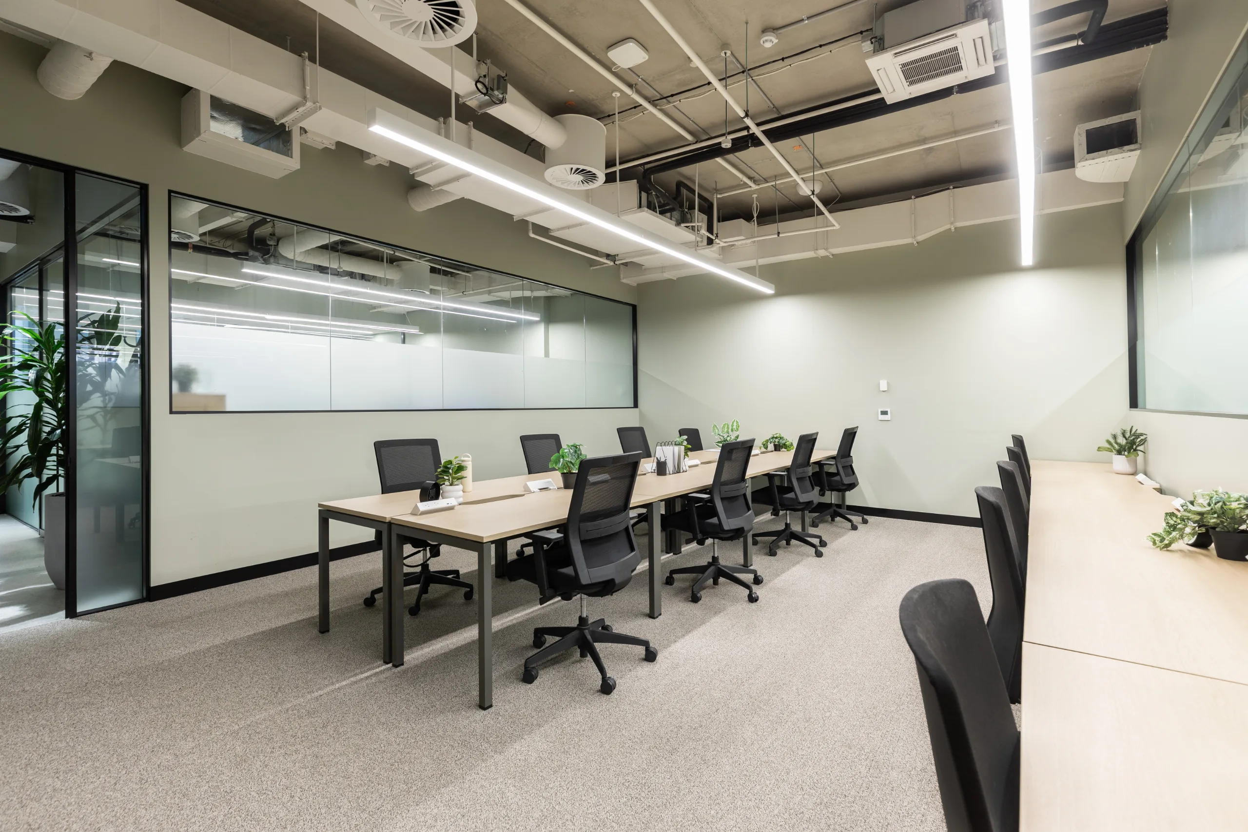 Bright, modern office space featuring long wooden desks, black ergonomic chairs, and large glass partitions.