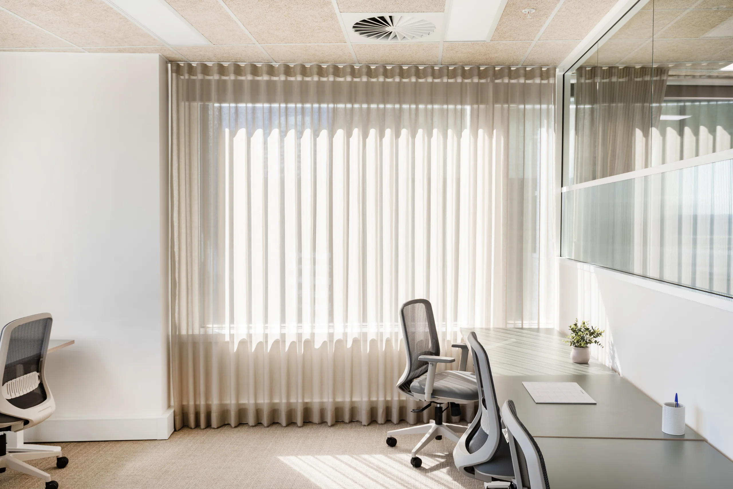 Bright office space featuring a long desk, ergonomic chairs, and large windows with sheer curtains.