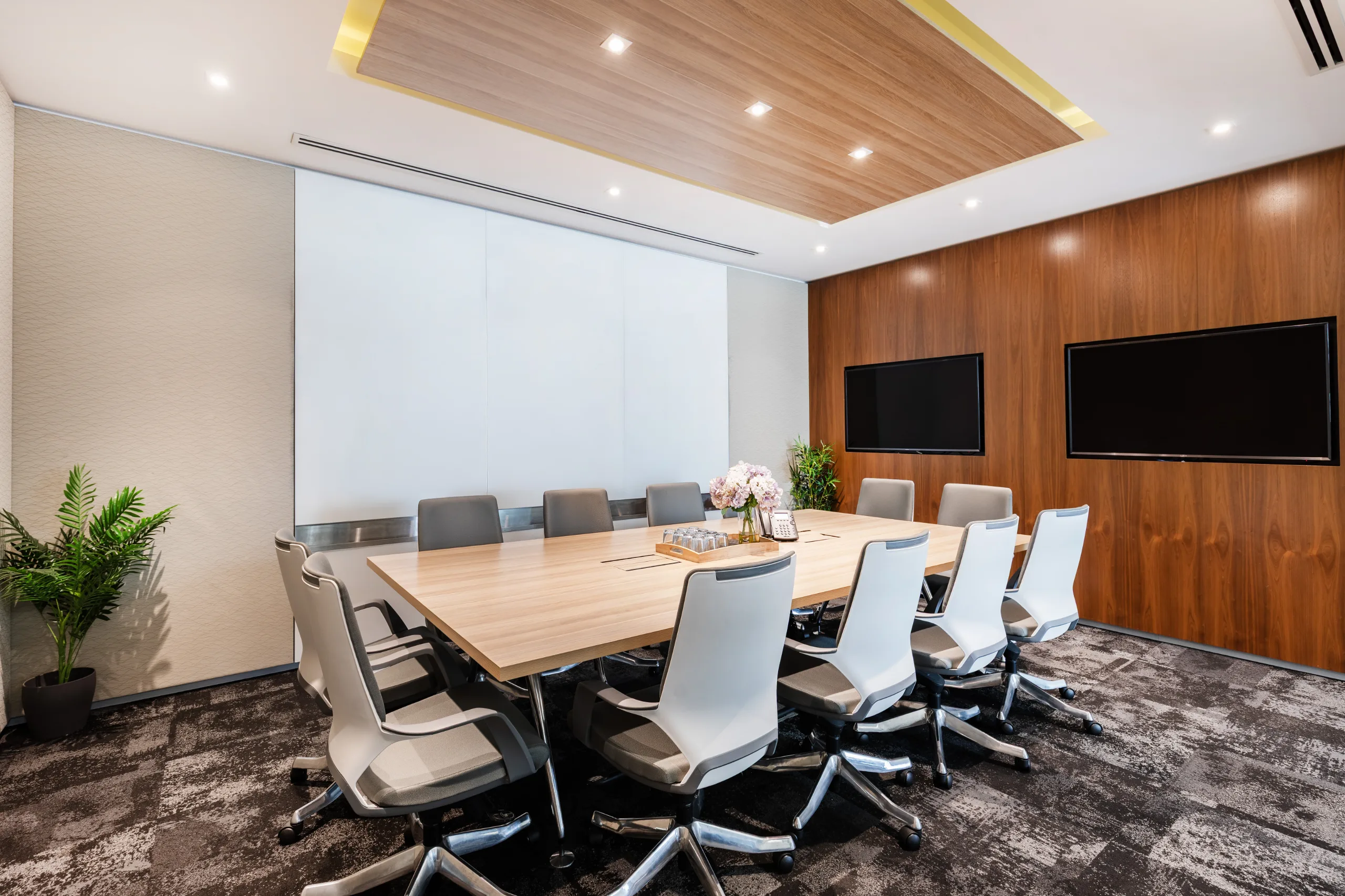 Modern conference room featuring a large wooden table, white chairs, and wall-mounted screens under bright recessed lighting.