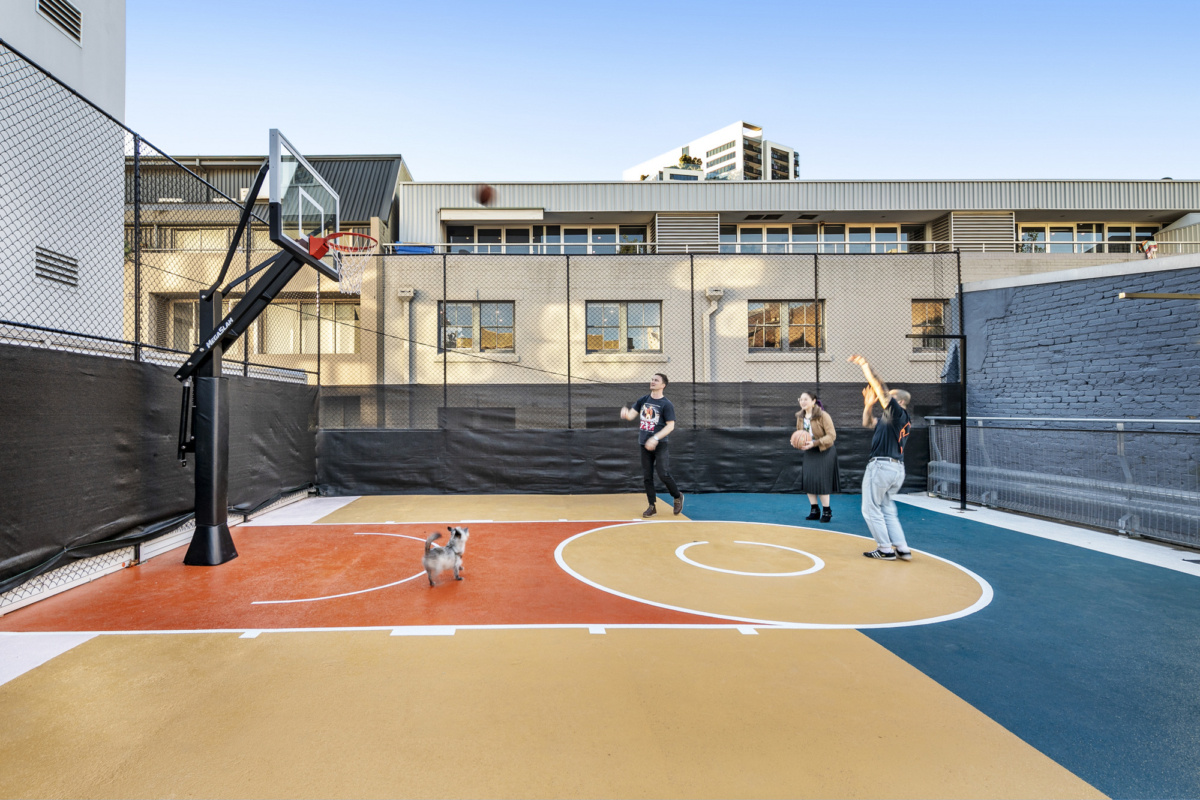 People play basketball on a colourful outdoor rooftop court with a small dog running nearby.