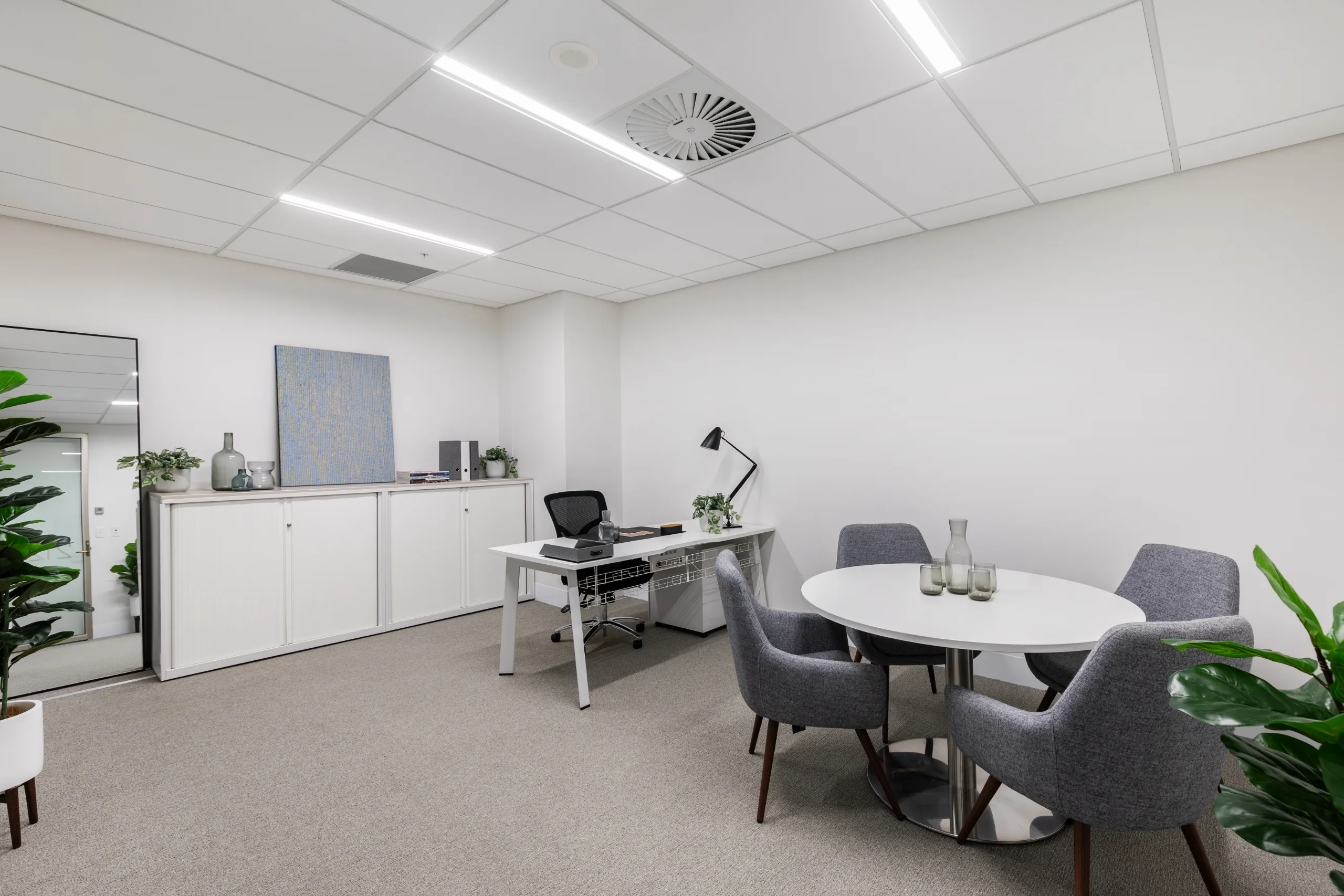 Brightly lit office space featuring a white desk, a round meeting table with grey chairs, and neutral carpeting.