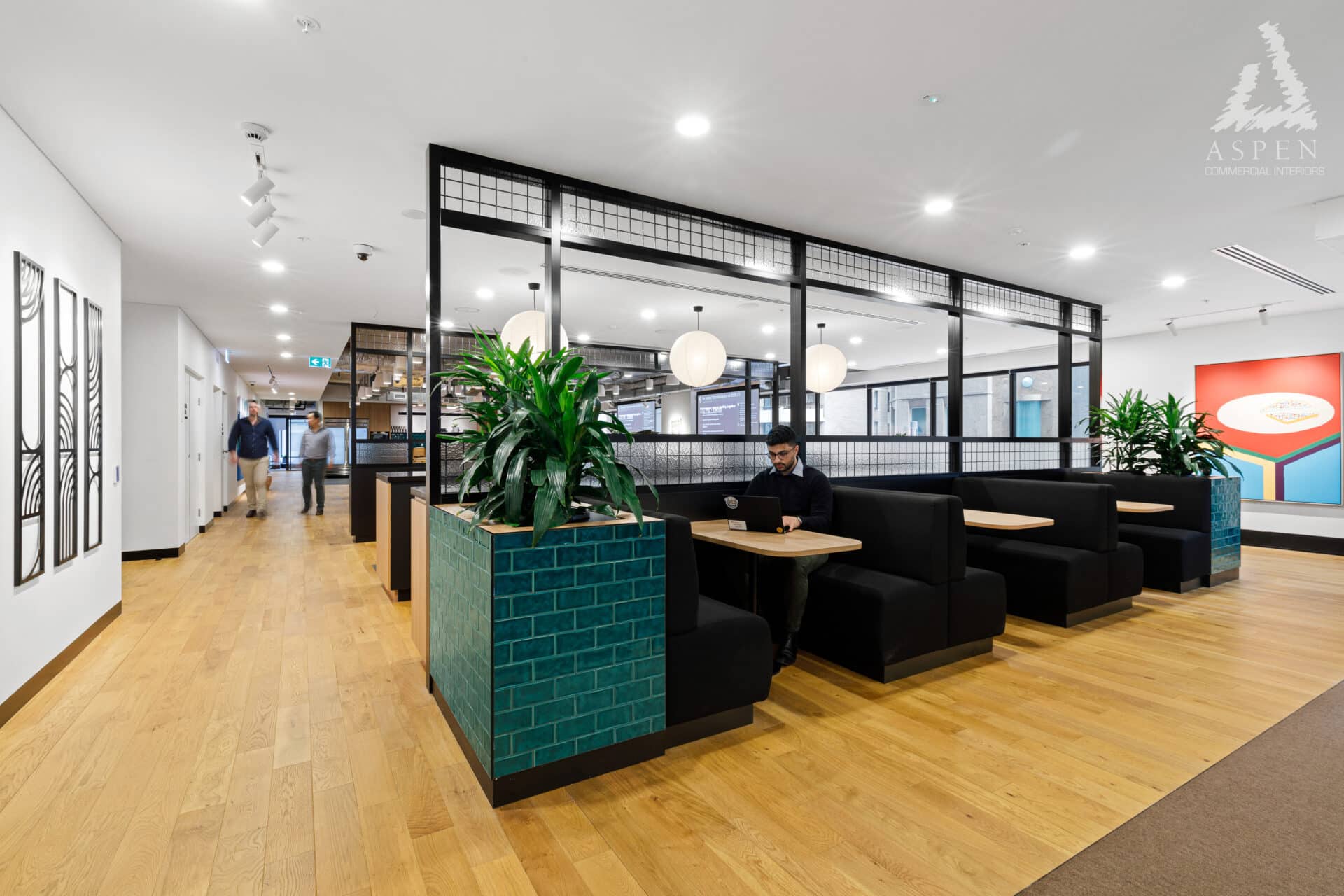 Modern office lounge with black booth seating, wooden floors, large green plants, and bright indoor lighting.