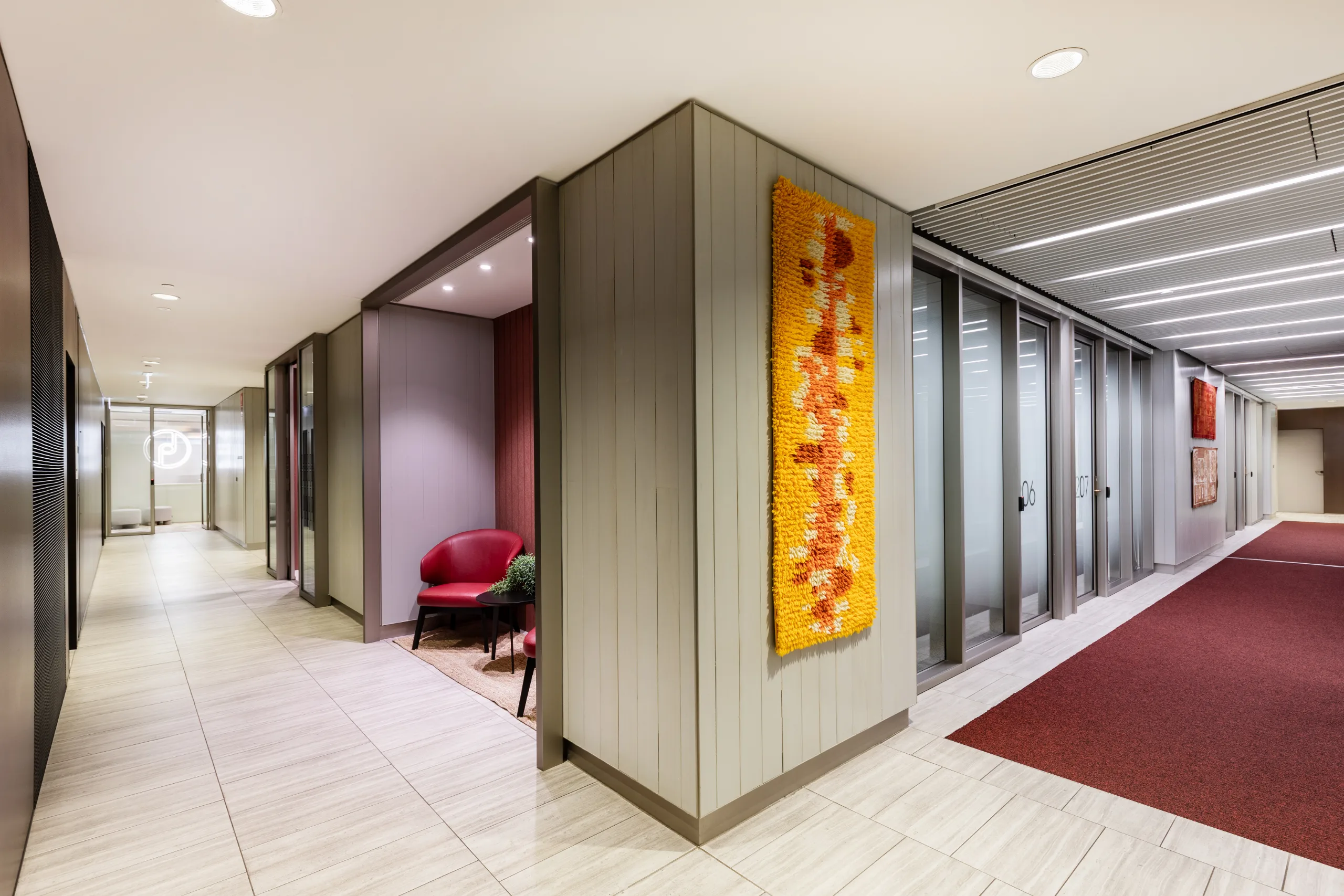 Bright office corridor featuring light tiled flooring, a red carpeted walkway, and a small seating area.